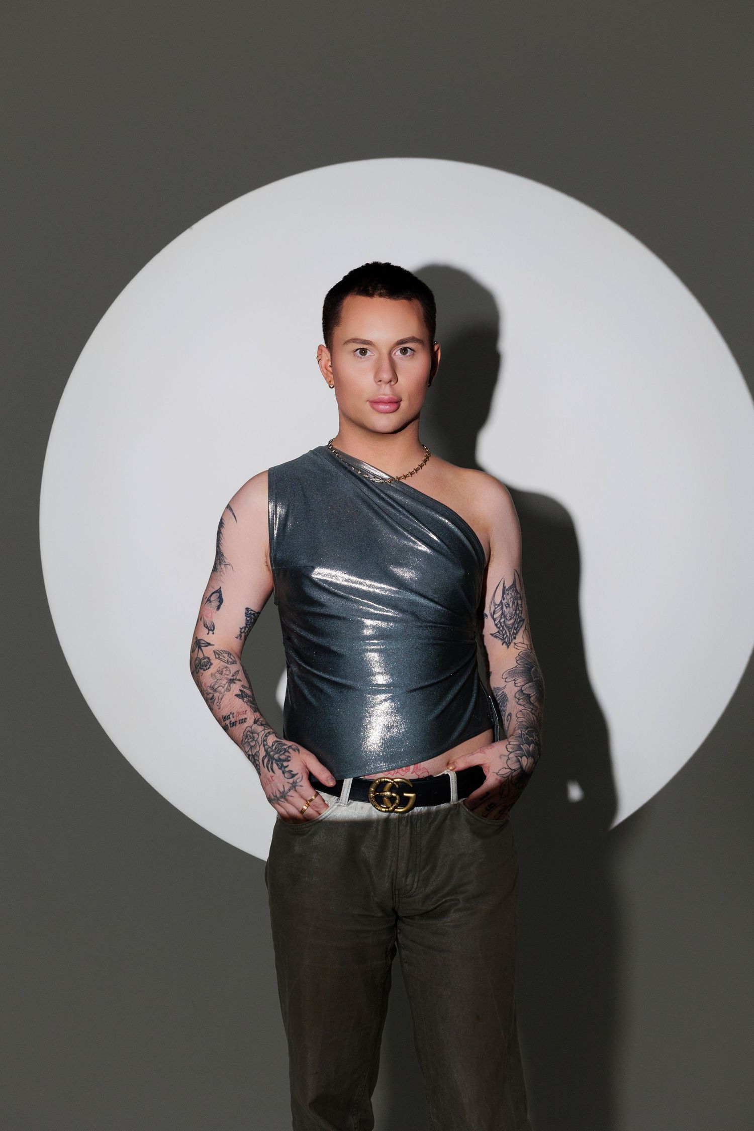Tattooed man in silver one-shoulder top and olive pants poses confidently in front of circular light backdrop.