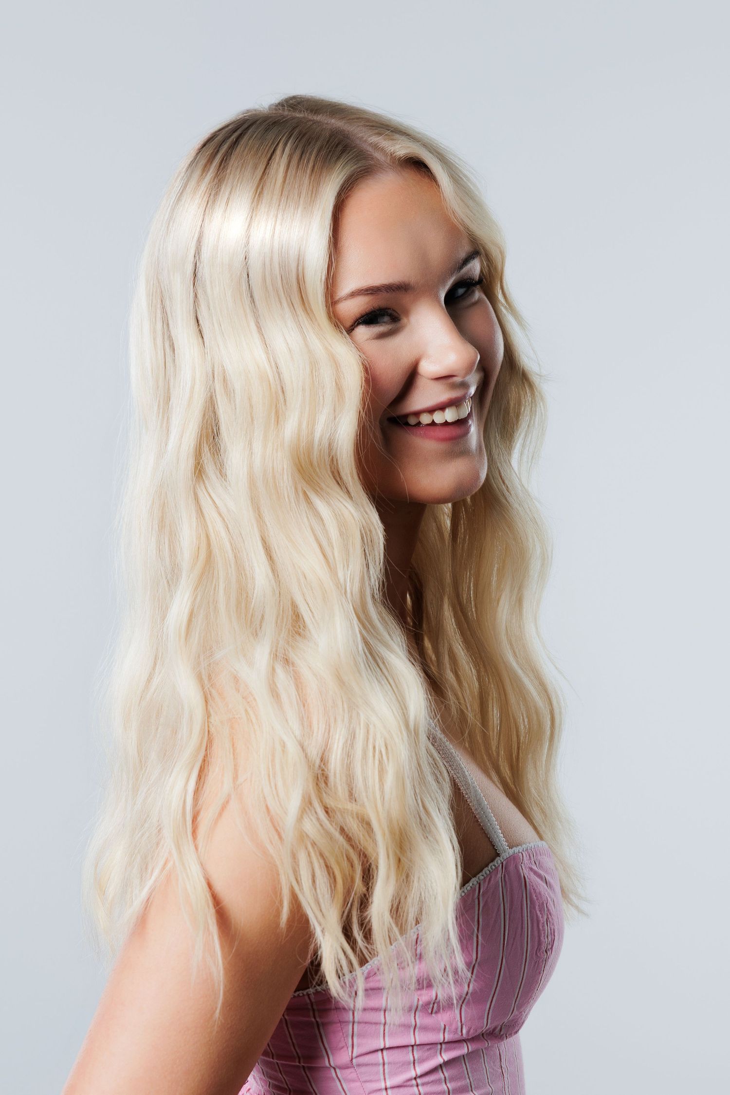 Smiling young woman with long wavy blonde hair wearing a pink top against a light gray background.