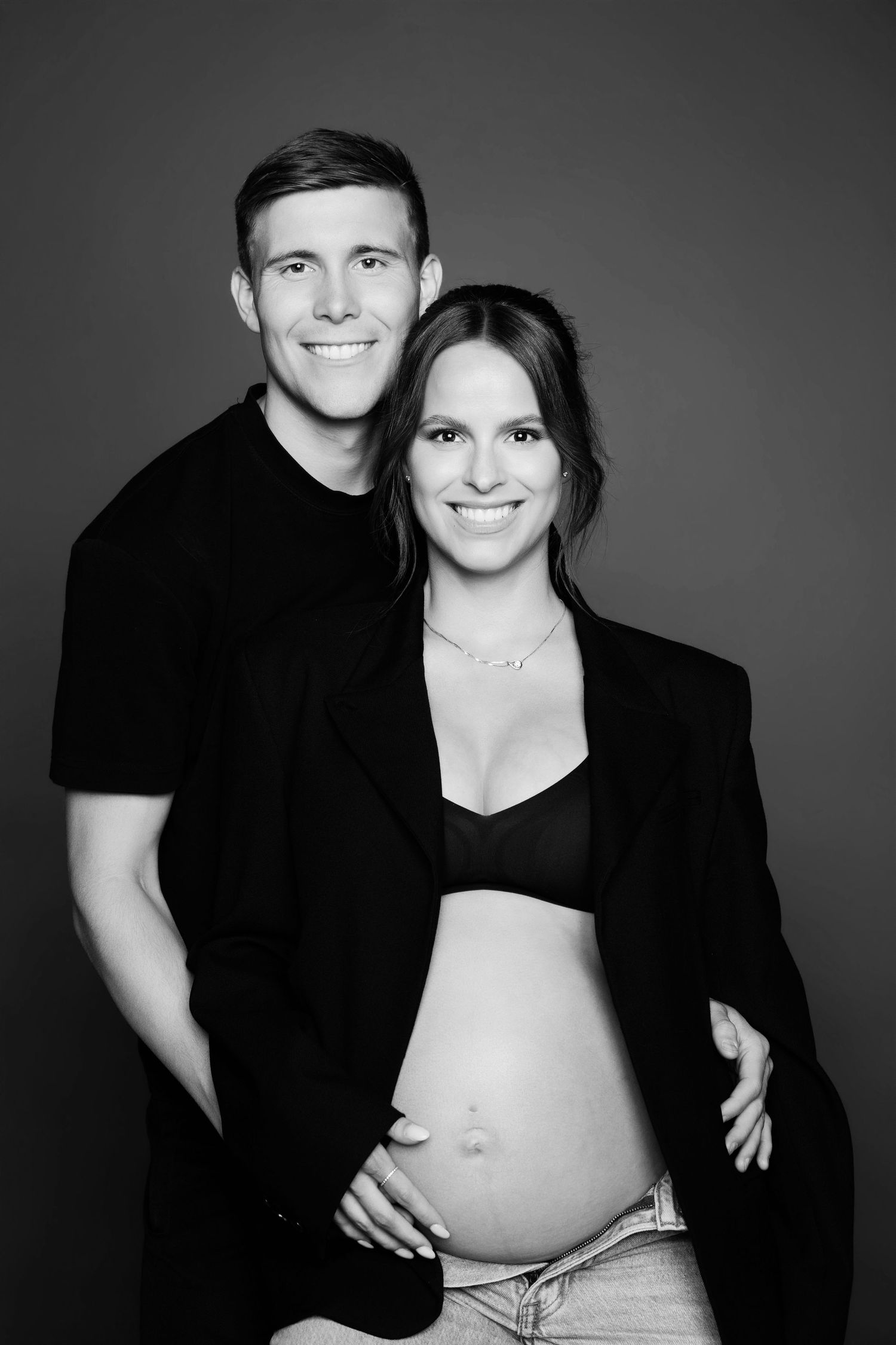 Black and white maternity photo of smiling couple, woman in black blazer showing pregnant belly.