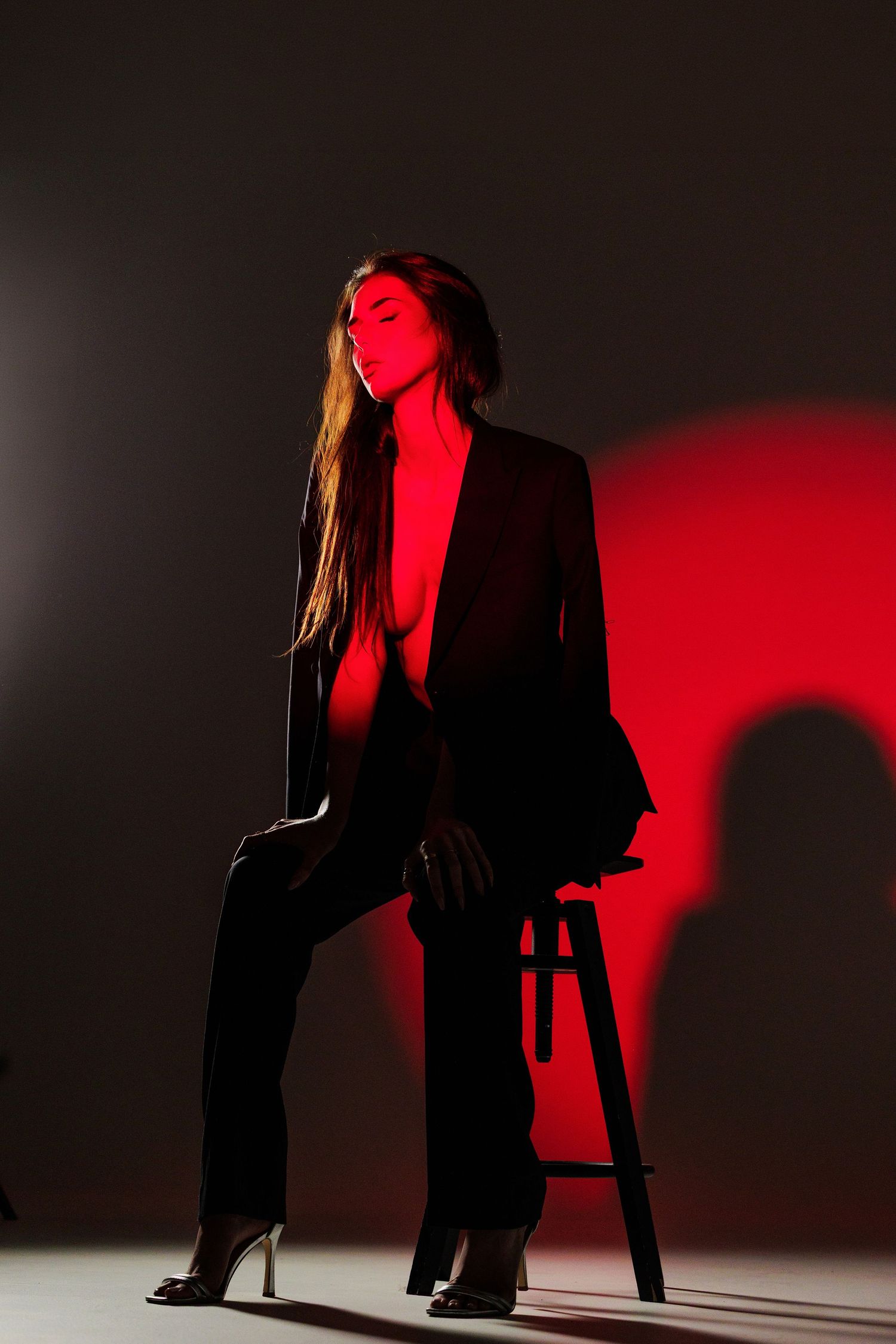 Woman in black suit seated on stool under dramatic red studio lighting, posing confidently with long hair.