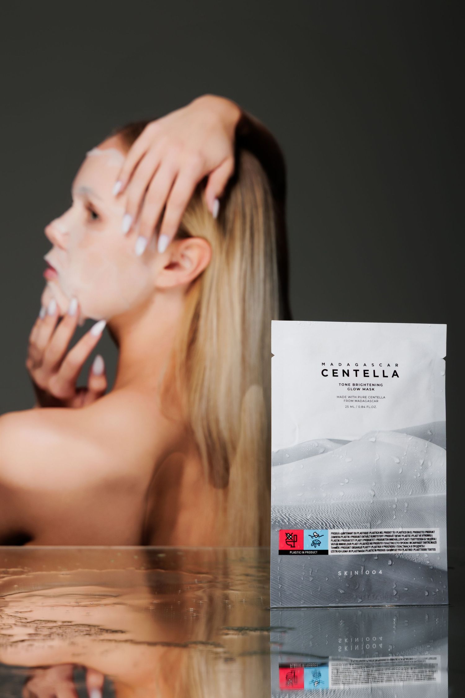 Madagascar Centella face mask packet displayed on reflective surface with blonde woman applying skincare in background.