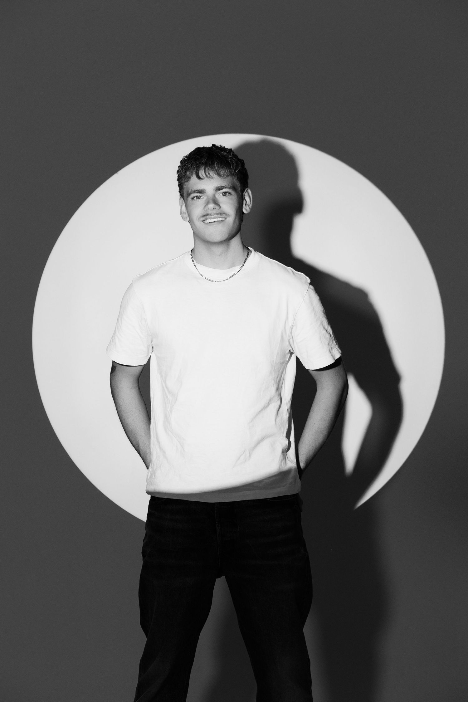 Young man in white t-shirt and black jeans poses confidently against a gray wall with circular white backdrop.