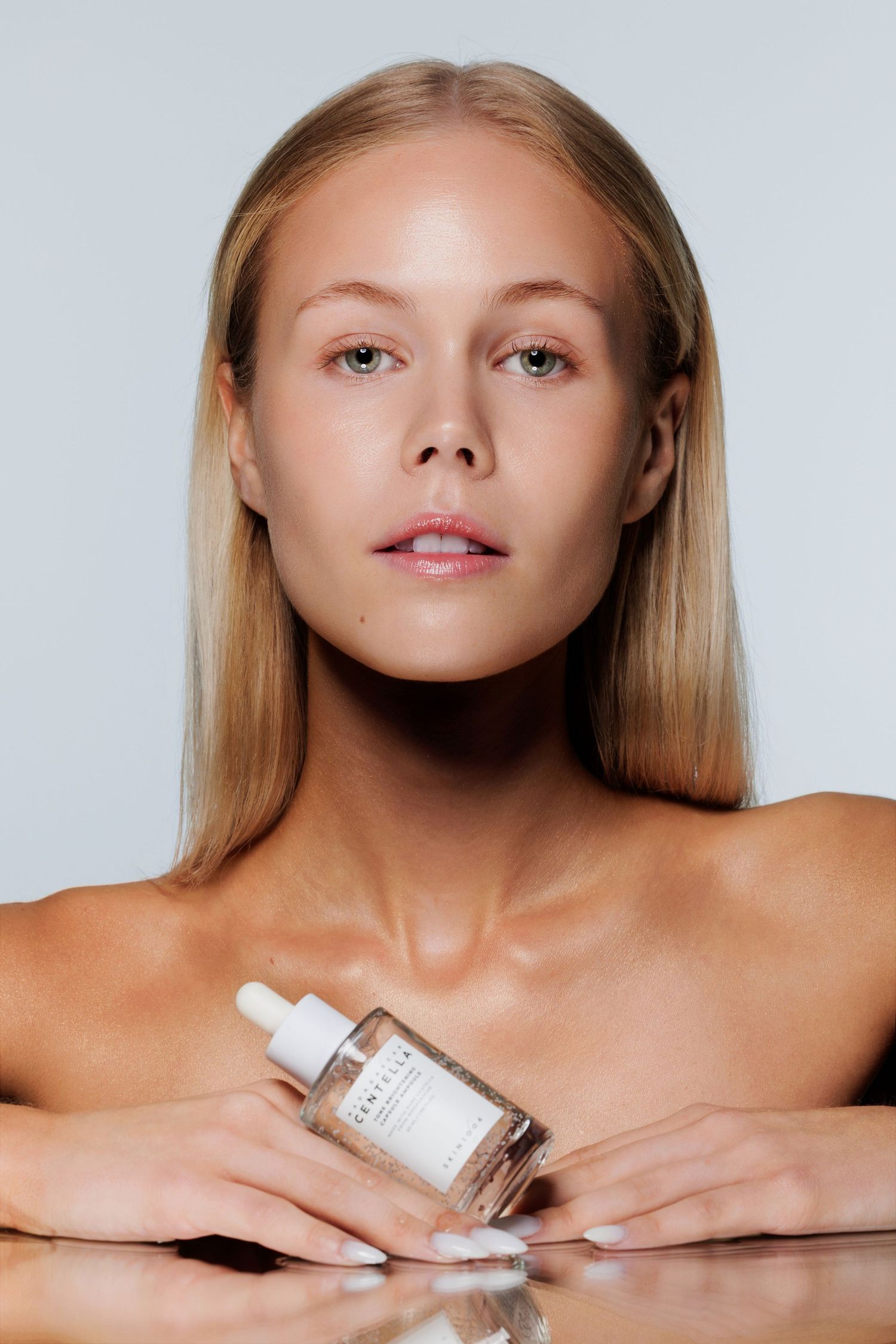Blonde model with natural makeup holding Centella serum bottle on mirrored table surface.