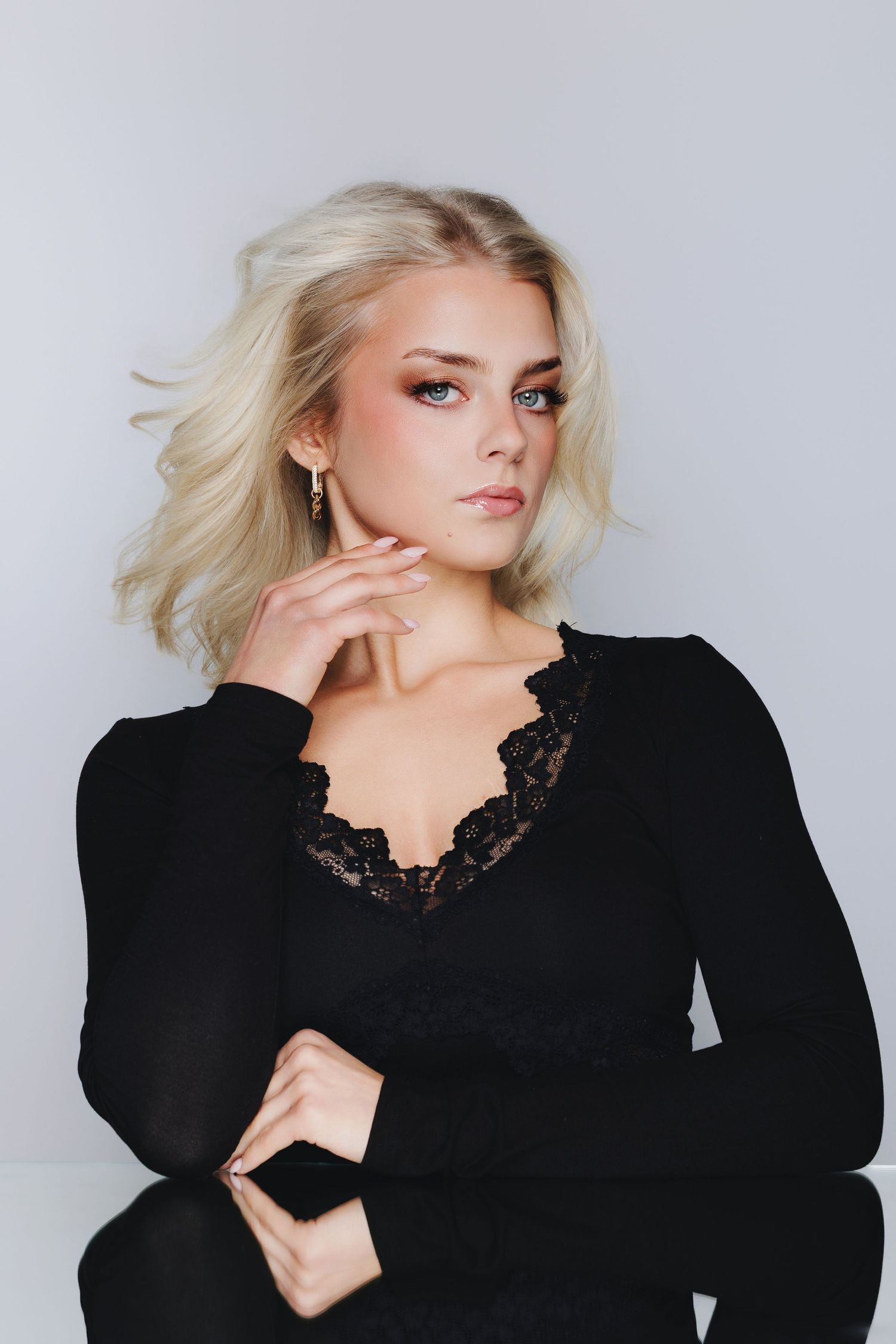 Blonde woman in black lace top poses thoughtfully against white background with smoky eye makeup.