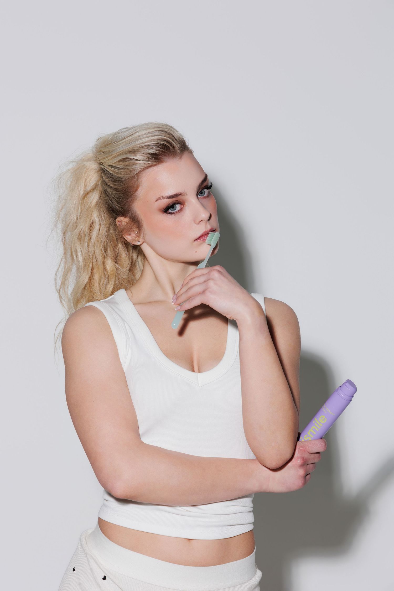 Blonde woman in white crop top holds purple object, posing thoughtfully against light gray background.