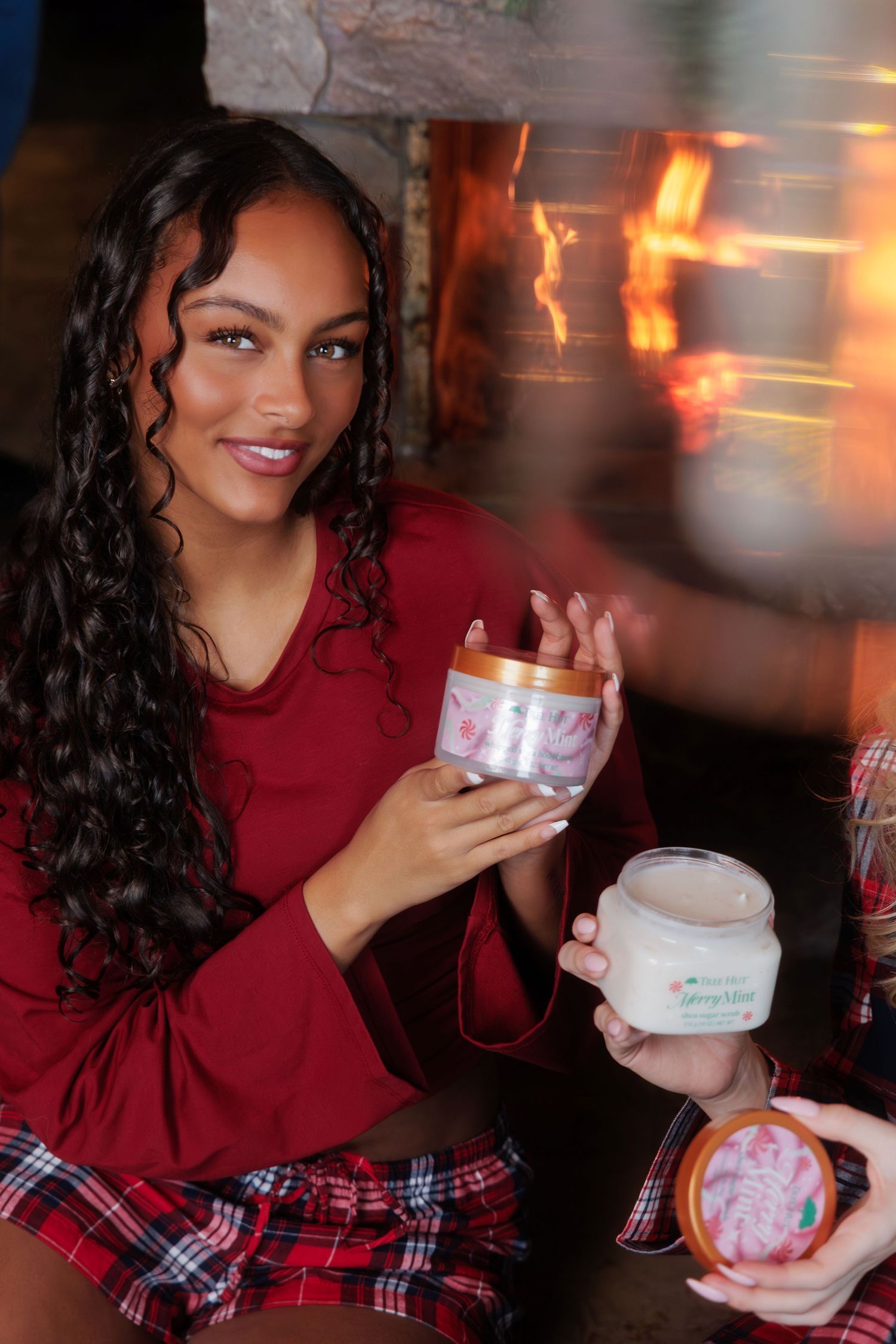 Woman in red pajamas holding Tree Hut Merry Mint sugar scrub jars by a cozy fireplace.