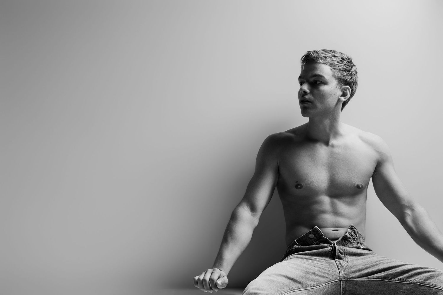 Black and white photo of muscular shirtless young man sitting against light gray wall.