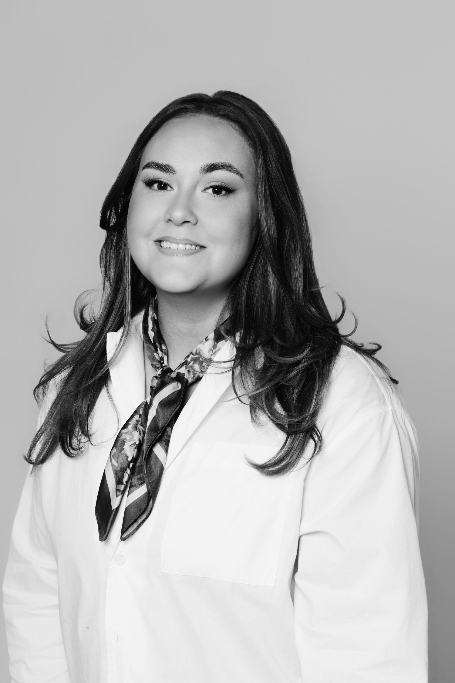 Black and white portrait of a smiling woman with long hair wearing a white blazer and patterned neck scarf.