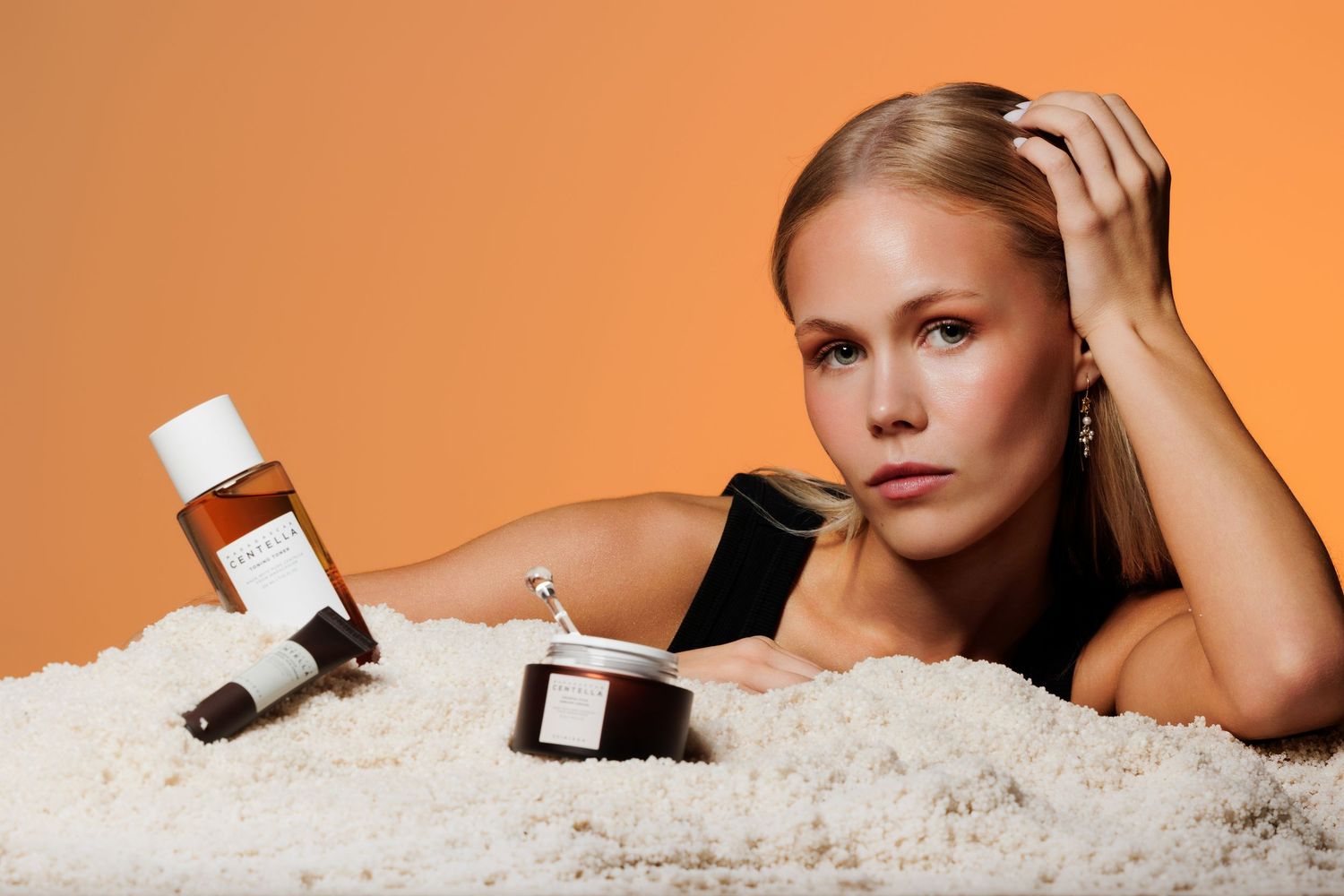 Blonde woman posing with luxury skincare products on white surface against orange background.