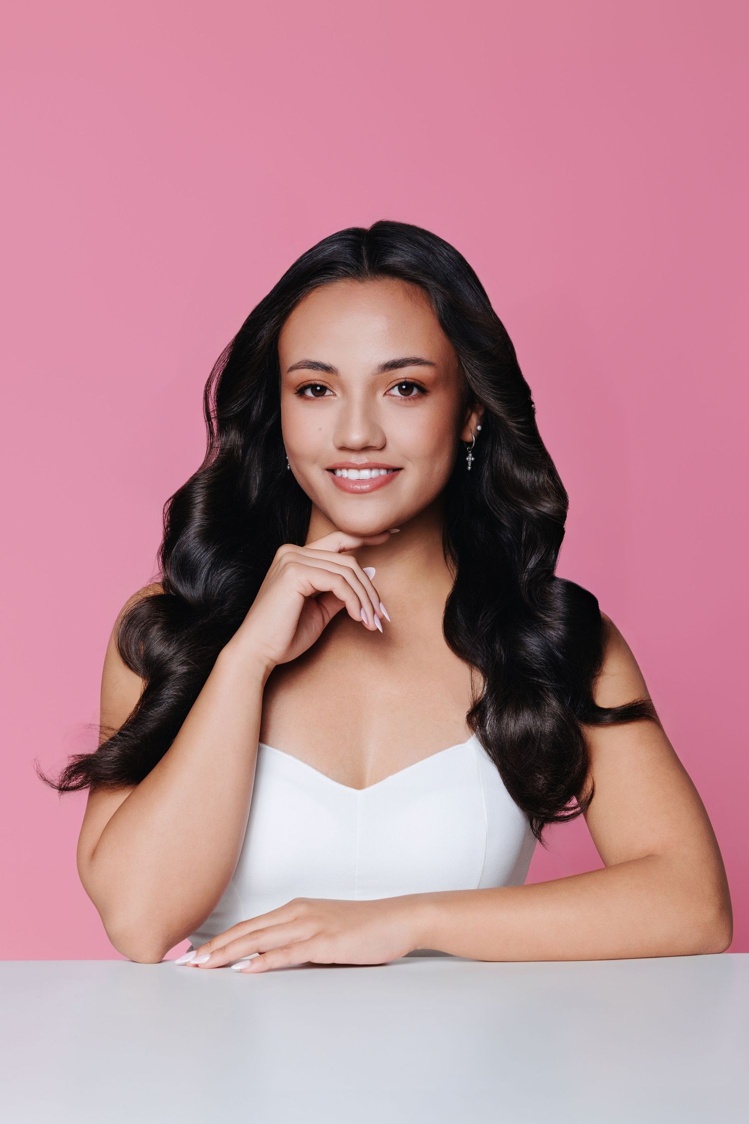 Young Asian woman with wavy dark hair in white top smiling at camera against pink background.
