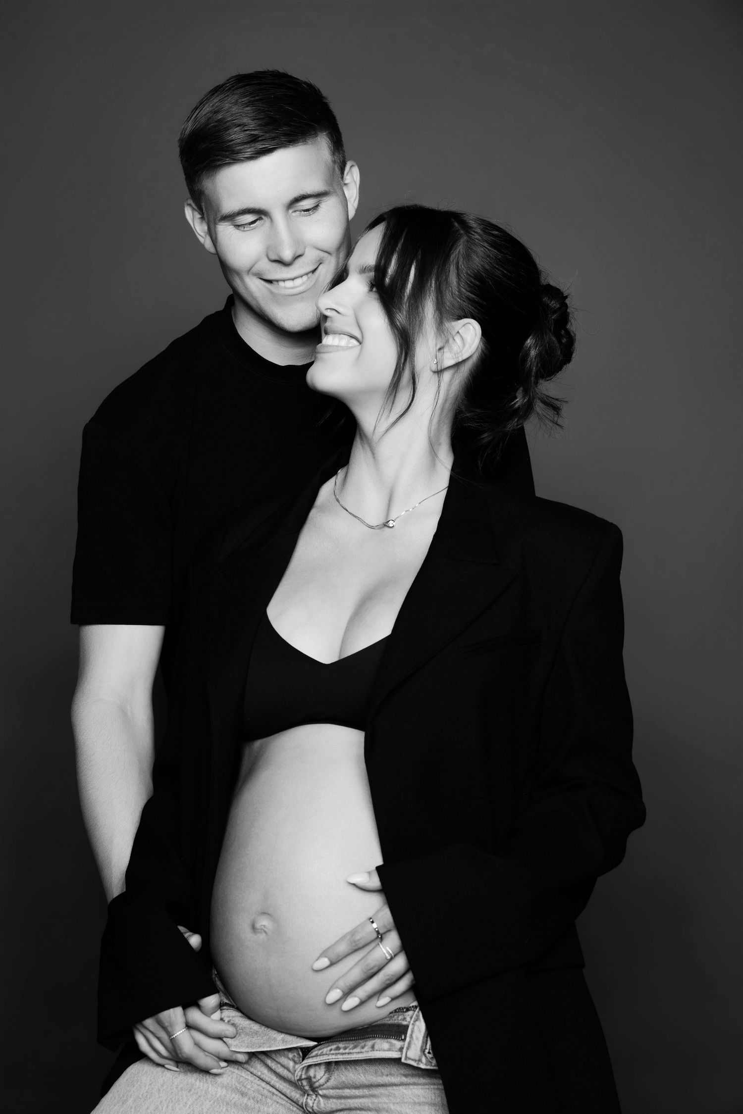 Black and white photo of smiling couple embracing, pregnant woman showing baby bump in studio.
