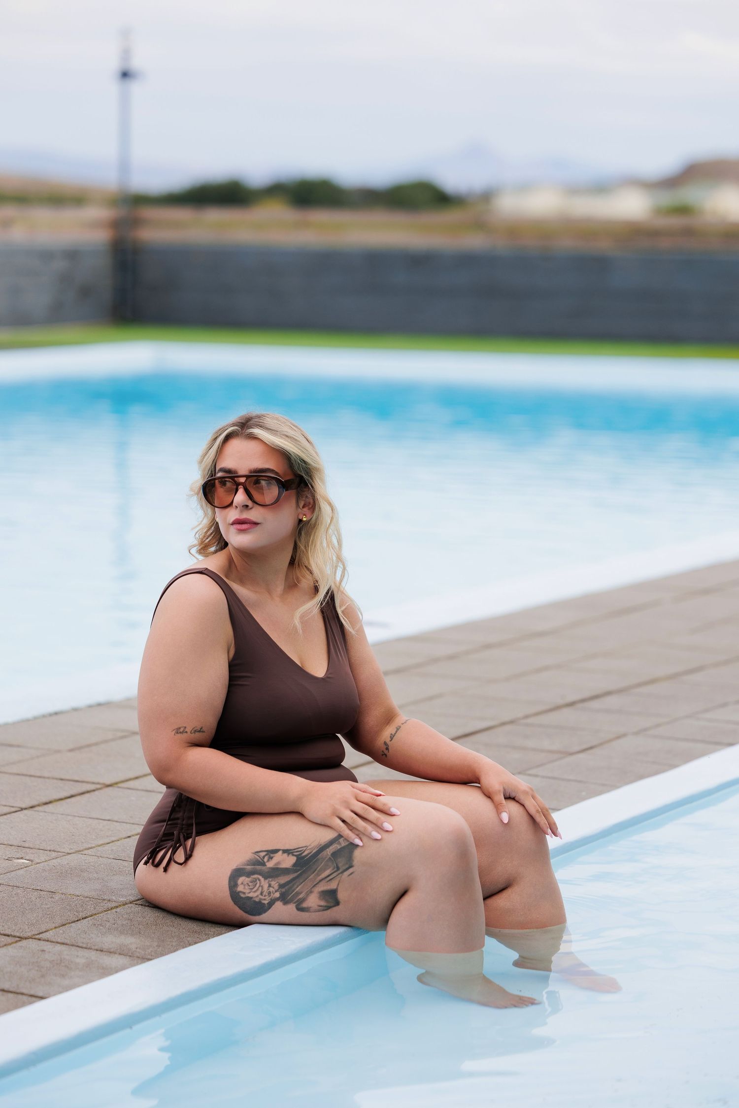 Plus-size blonde woman in brown swimsuit with tattoos sitting poolside wearing sunglasses outdoors.