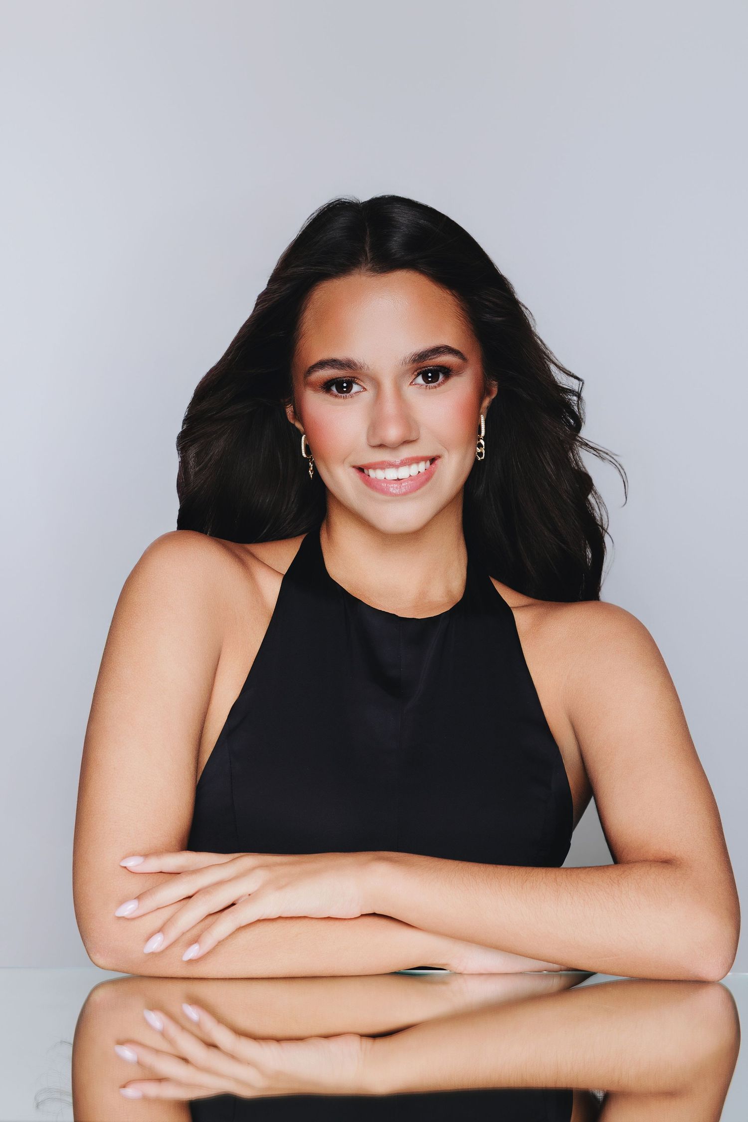 Young woman with dark hair smiling, wearing a black halter top, seated at a reflective table against a gray background.