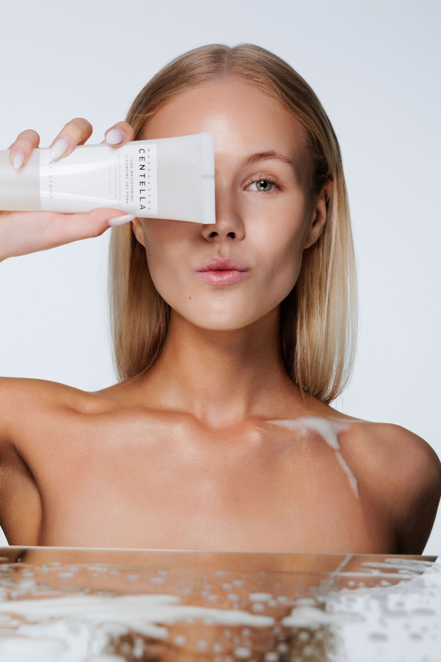 Woman holding Centella skincare tube over her eye, with water reflection, against white background.