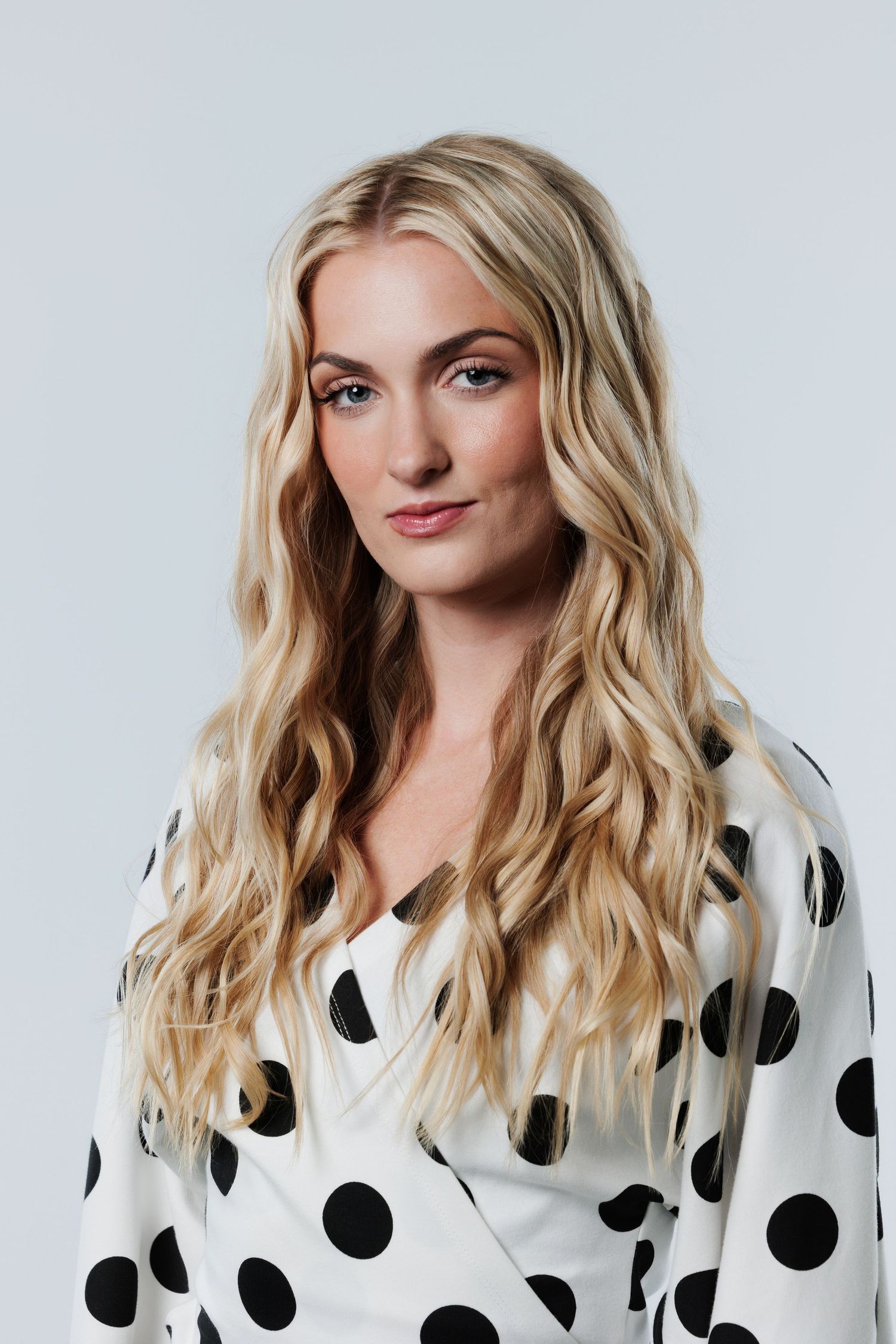 Blonde woman with long wavy hair wearing a white and black polka dot wrap blouse, posing against a light gray background.