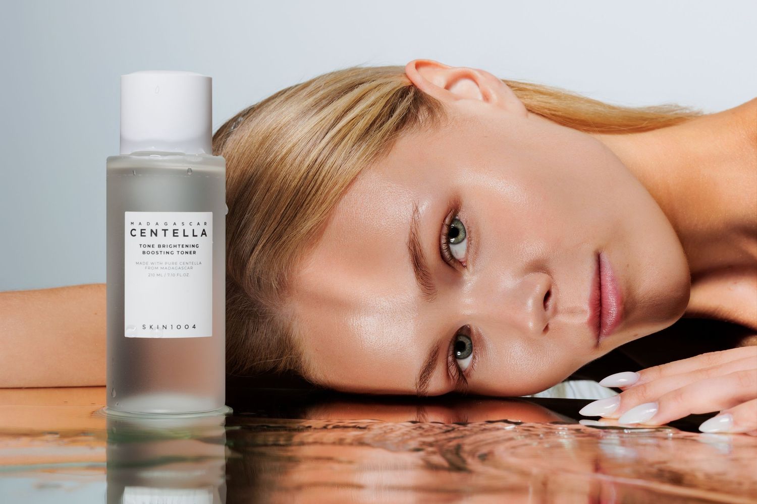 Woman resting beside a SKIN1004 Centella toner bottle on a reflective surface, showcasing the skincare product.
