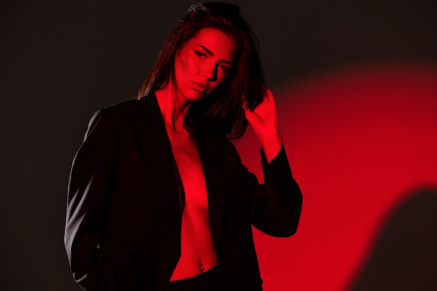 Woman in black blazer poses dramatically under red and dark studio lighting in a moody fashion portrait.