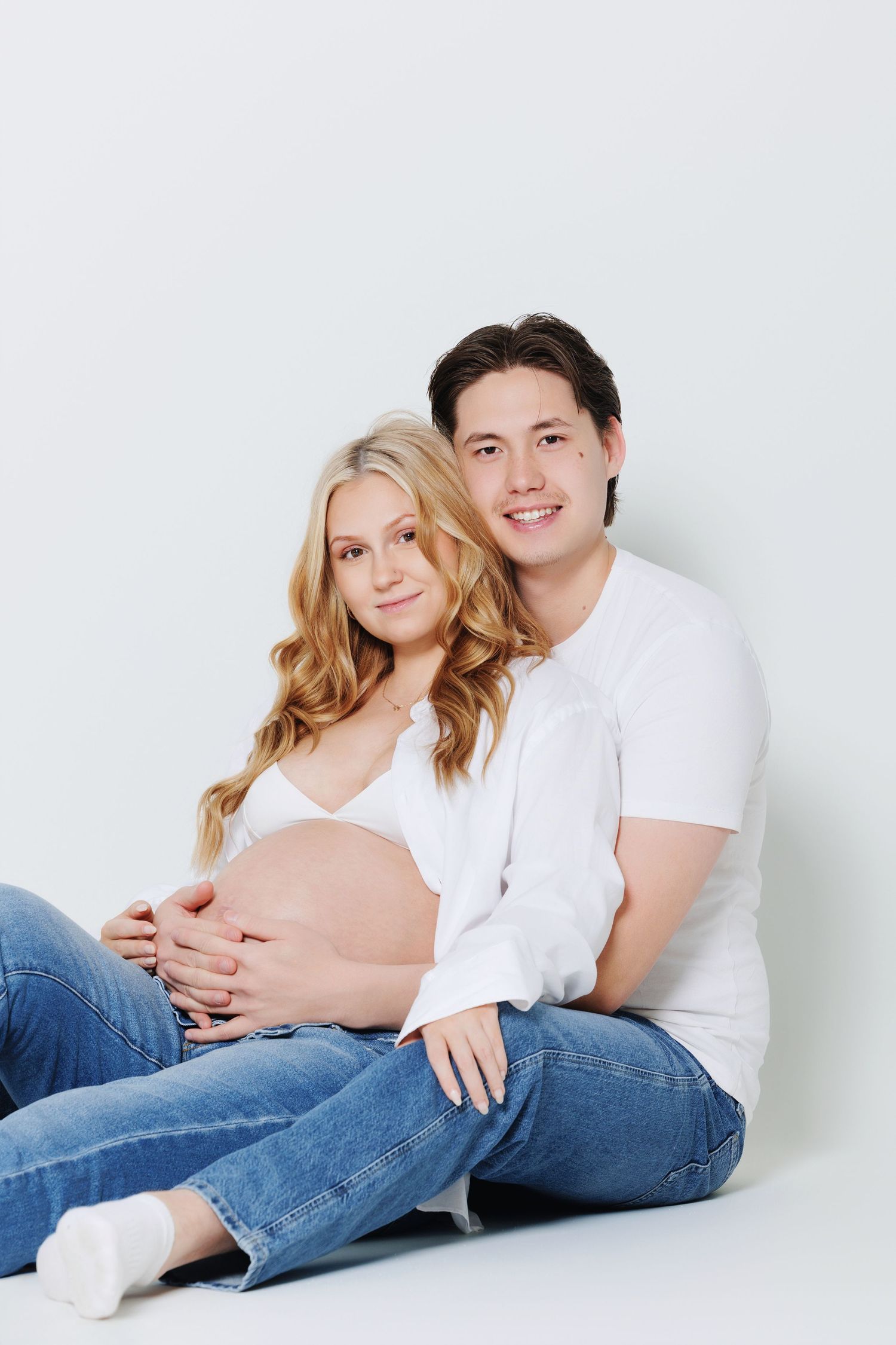 Happy couple sitting on floor, pregnant blonde woman and smiling man holding her baby bump.