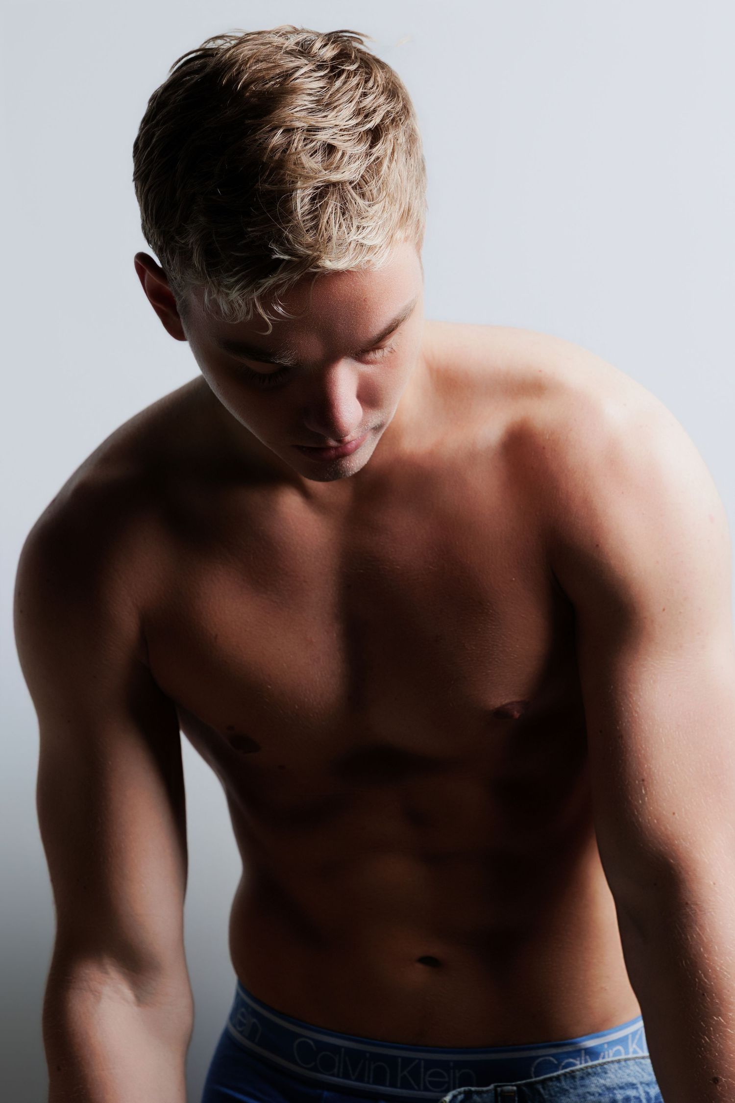 Muscular shirtless blonde man looking down in dramatic studio lighting wearing blue jeans.