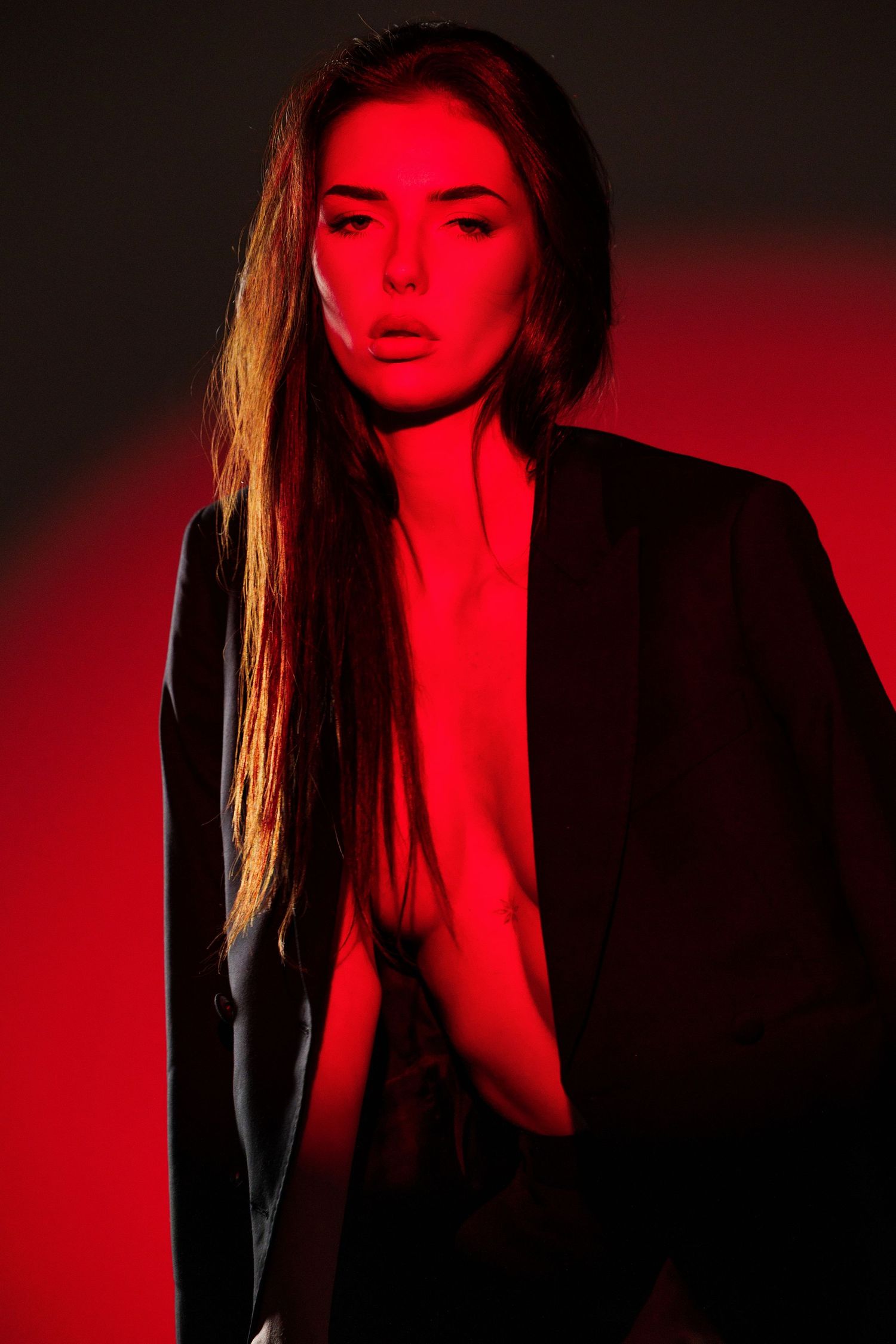 Woman in black blazer posed against red-lit background with dramatic lighting creating moody atmosphere.