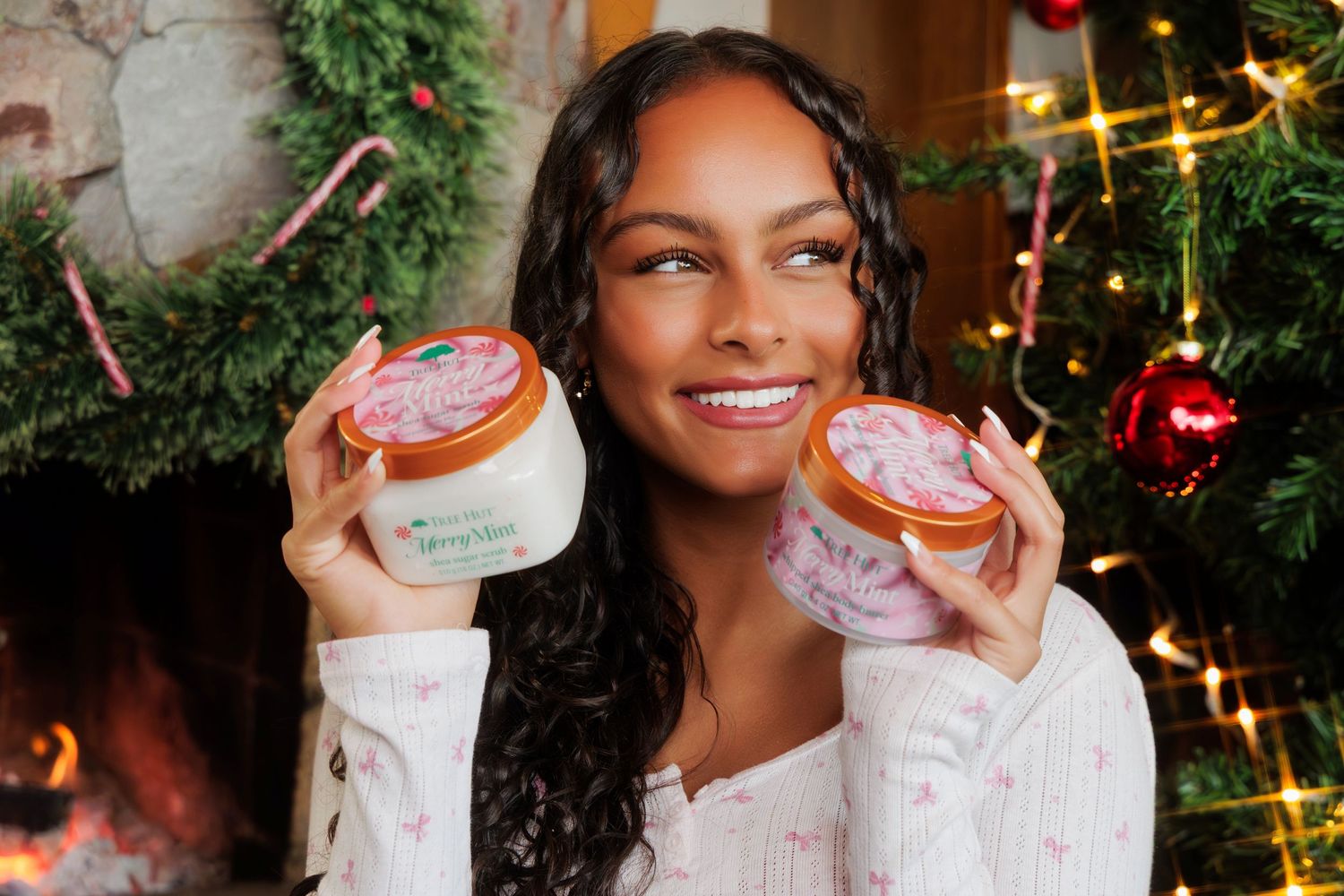 Smiling woman holding two Tree Hut Merry Mint sugar scrub jars near a Christmas tree.