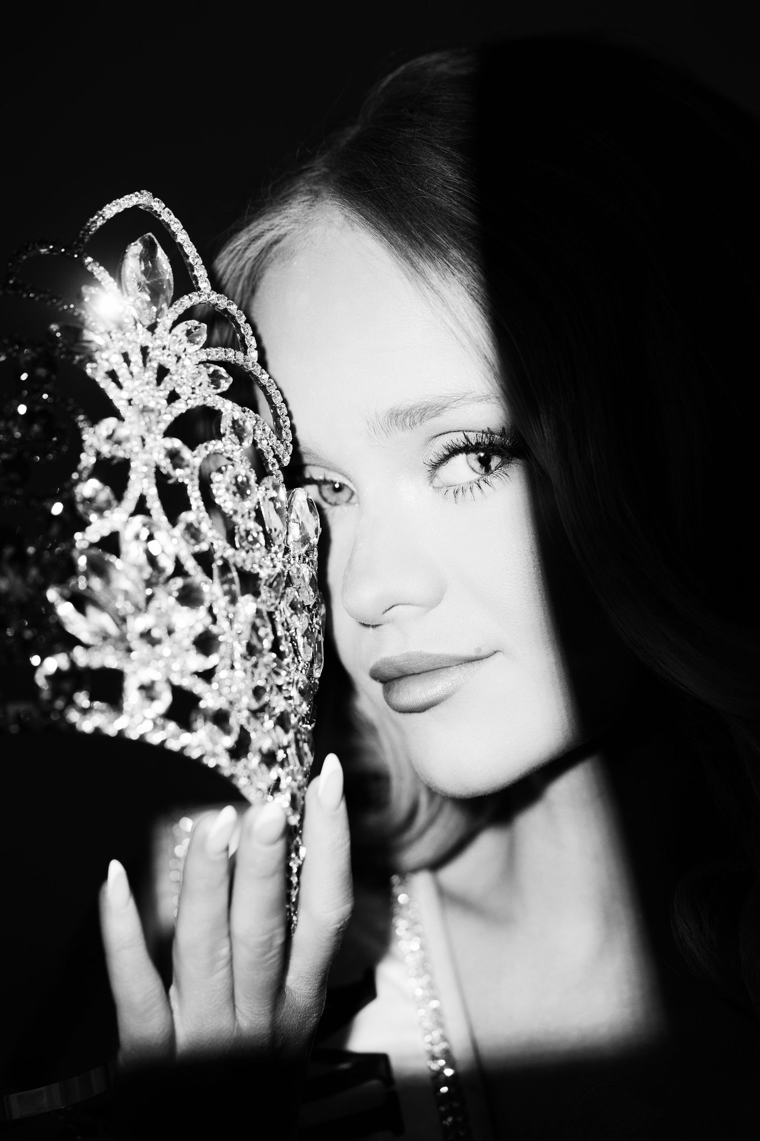 Black and white portrait of a young woman holding a sparkling diamond tiara crown next to her face.