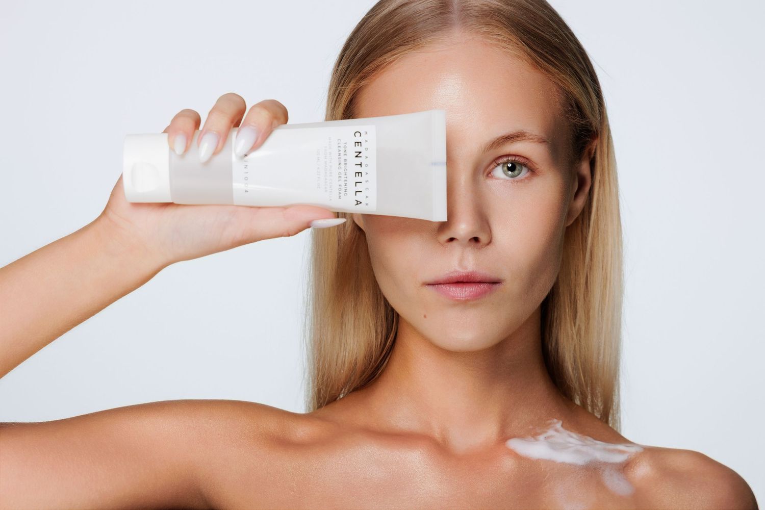 Woman holding a white Centella skincare bottle over her eye against a light background, showcasing clean beauty product.