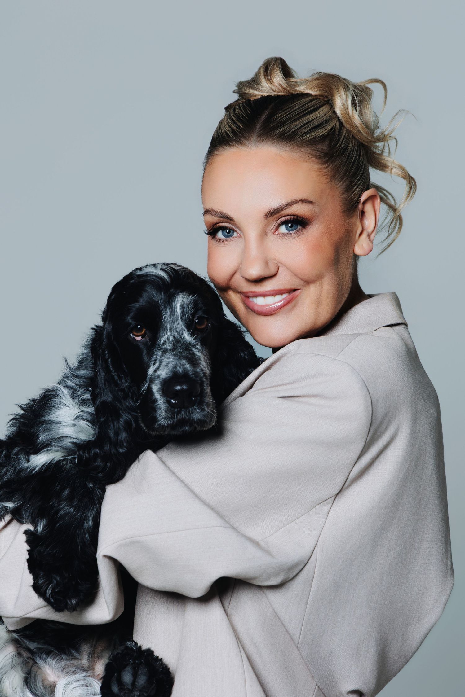 Smiling blonde woman with blue eyes holding a black and white Cocker Spaniel dog against a gray background.