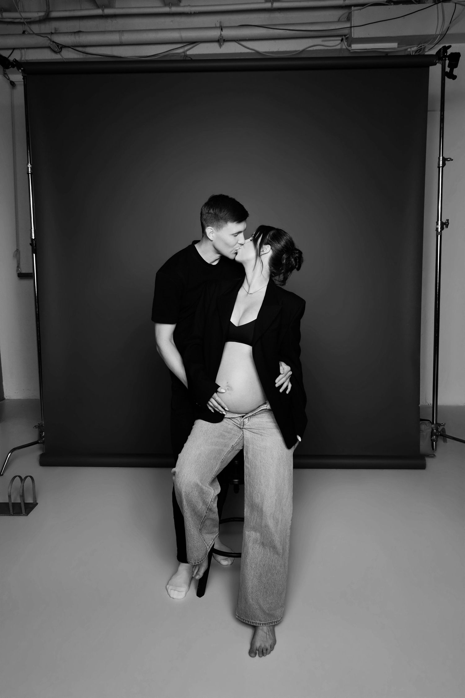 Black and white photo of couple kissing, pregnant woman seated showing bare belly in photo studio.