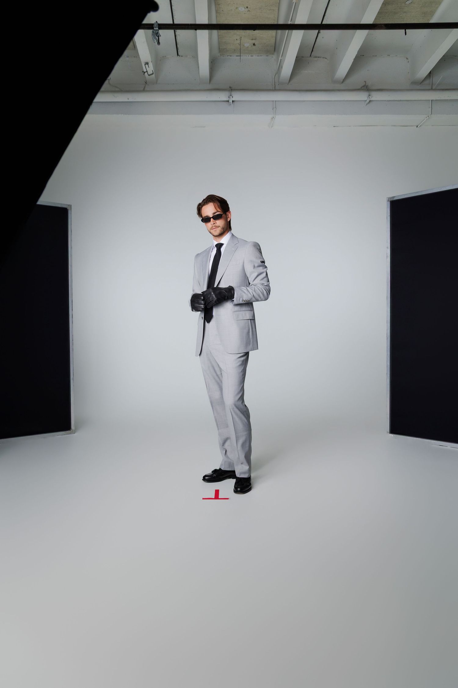 Man in gray suit with black leather gloves and sunglasses standing in photo studio.