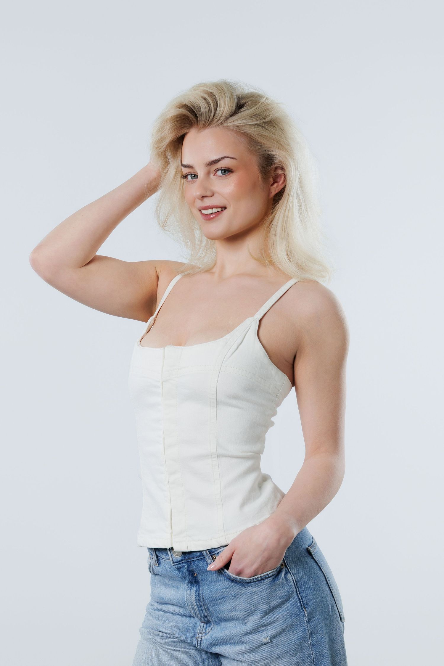Smiling blonde woman in white camisole top and blue jeans posing against light gray background.
