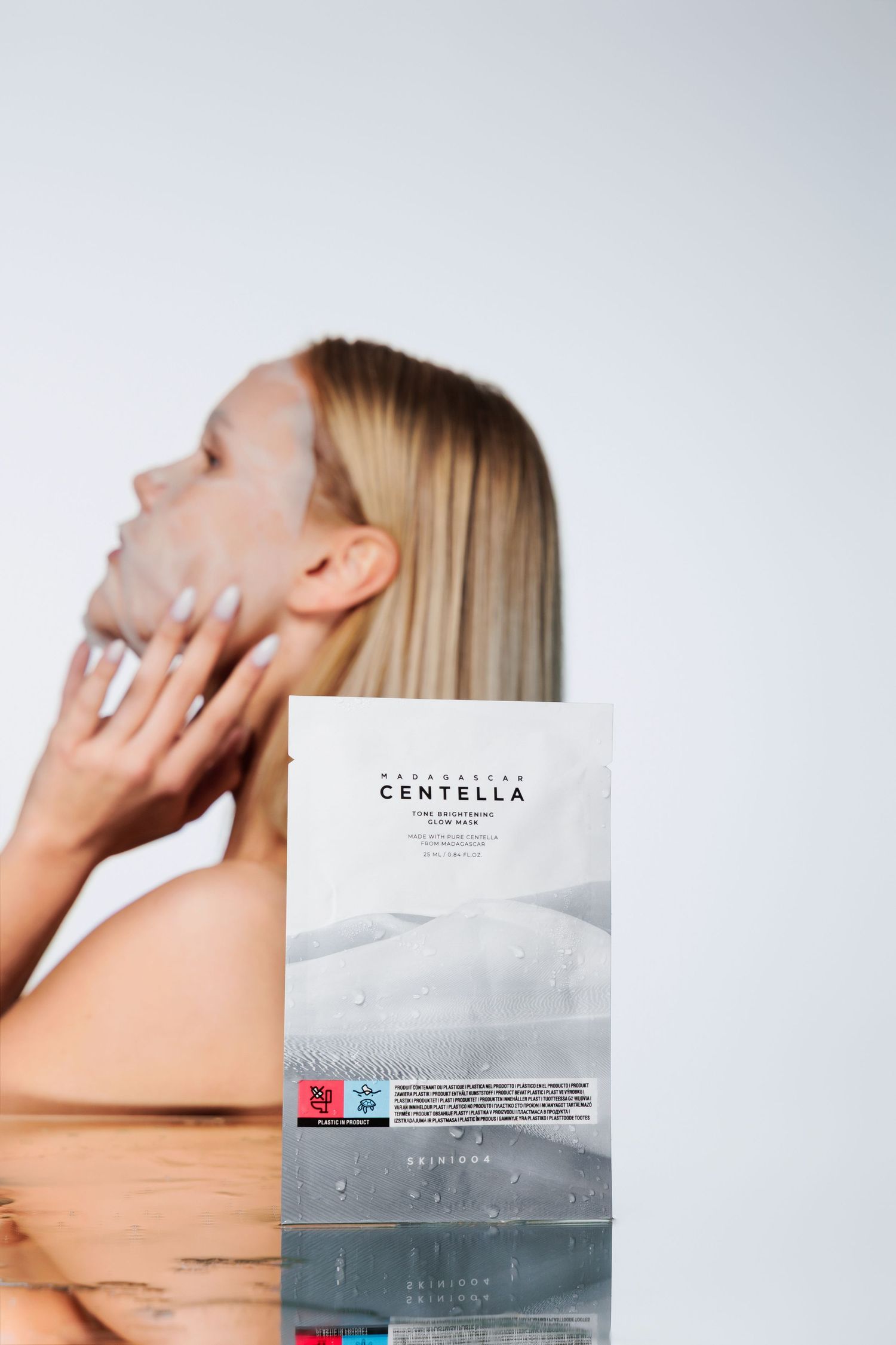 Woman applying Centella skin brightening face mask with product box in foreground on white background.