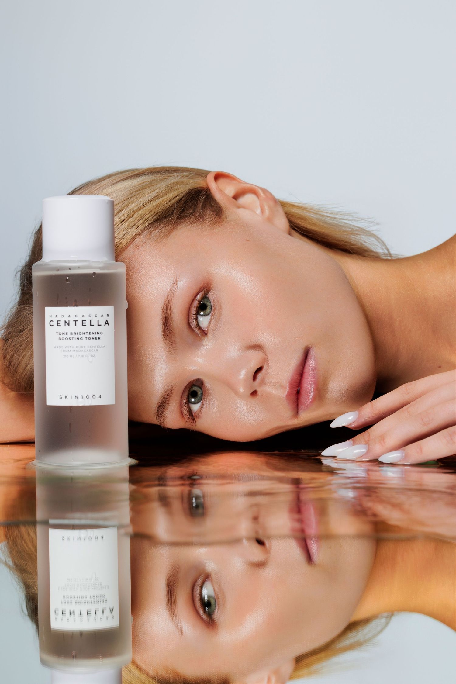 Blonde woman resting face on mirror surface beside Centella soothing toner bottle.