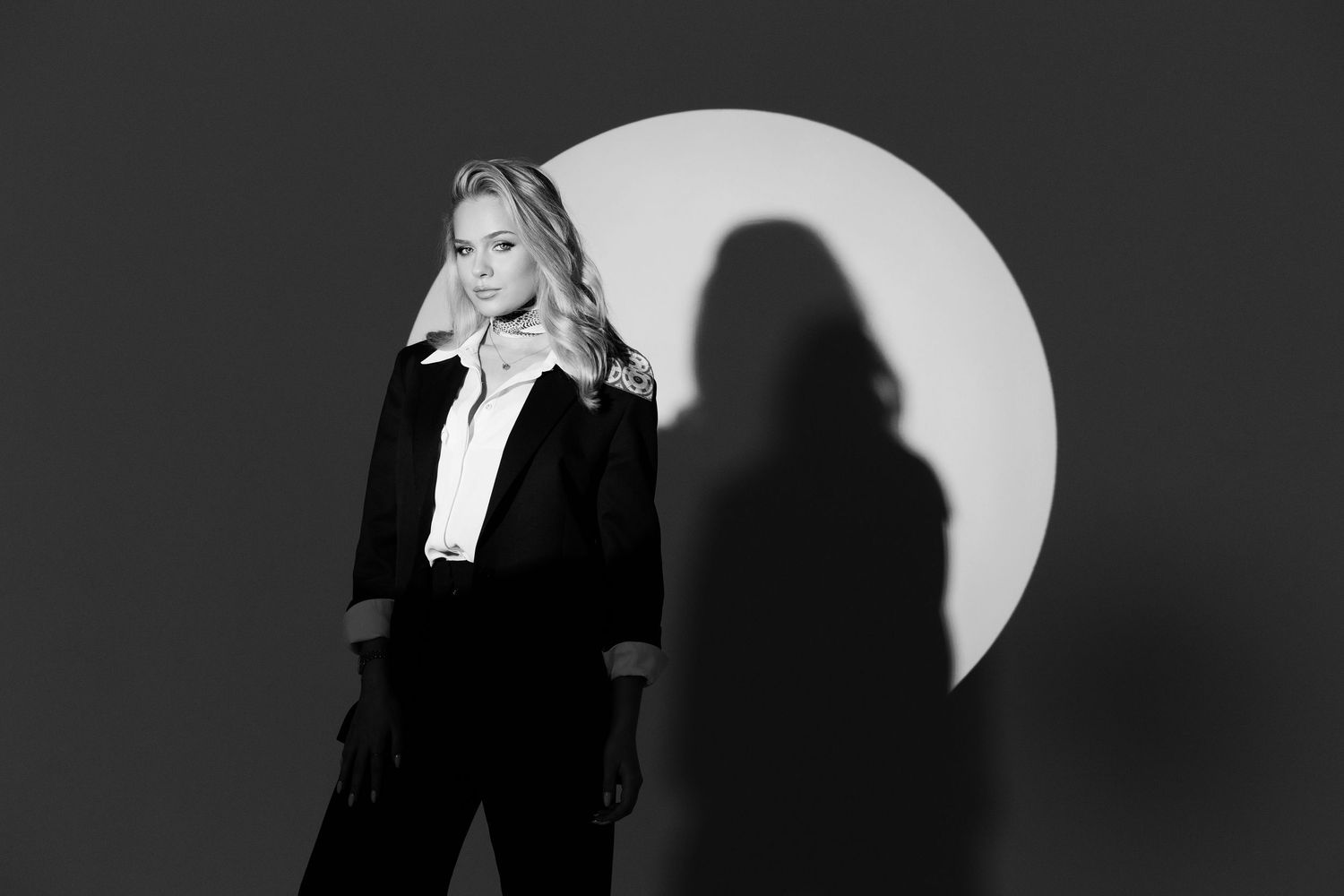 Black and white photo of a blonde woman in a black suit posing confidently in front of a circular light backdrop.