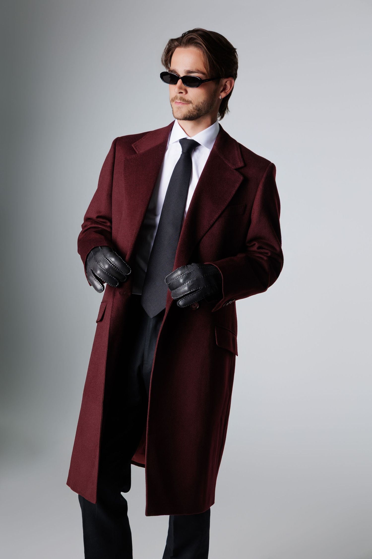 Man in burgundy overcoat with black tie and leather gloves posing on gray background.