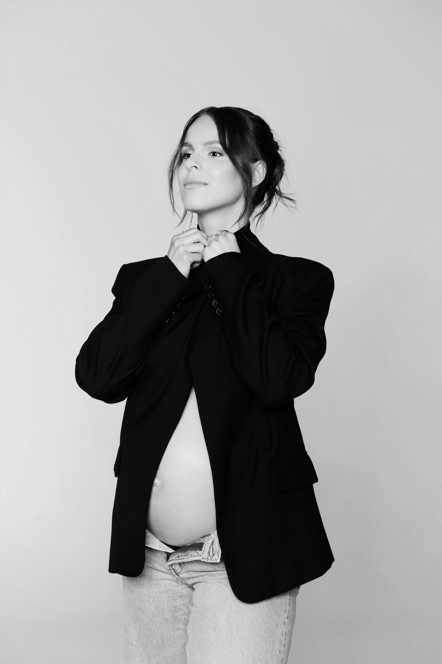 Black and white photo of pregnant woman in black blazer and jeans looking up thoughtfully in studio.