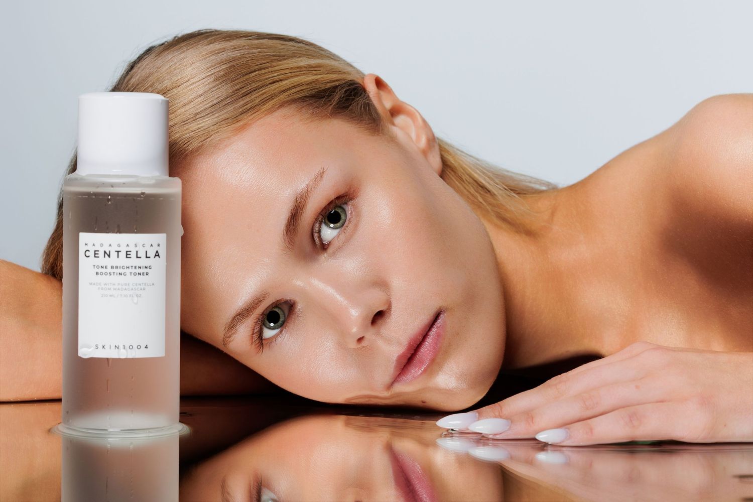 Woman resting her chin on a reflective surface next to a Centella toner bottle in a minimalist skincare product shot.