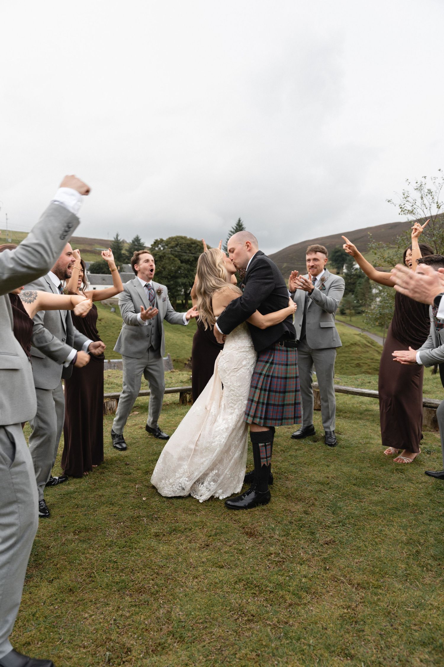 Bride and groom sharing first kiss surrounded by cheering wedding party on grassy Scottish hillside.
