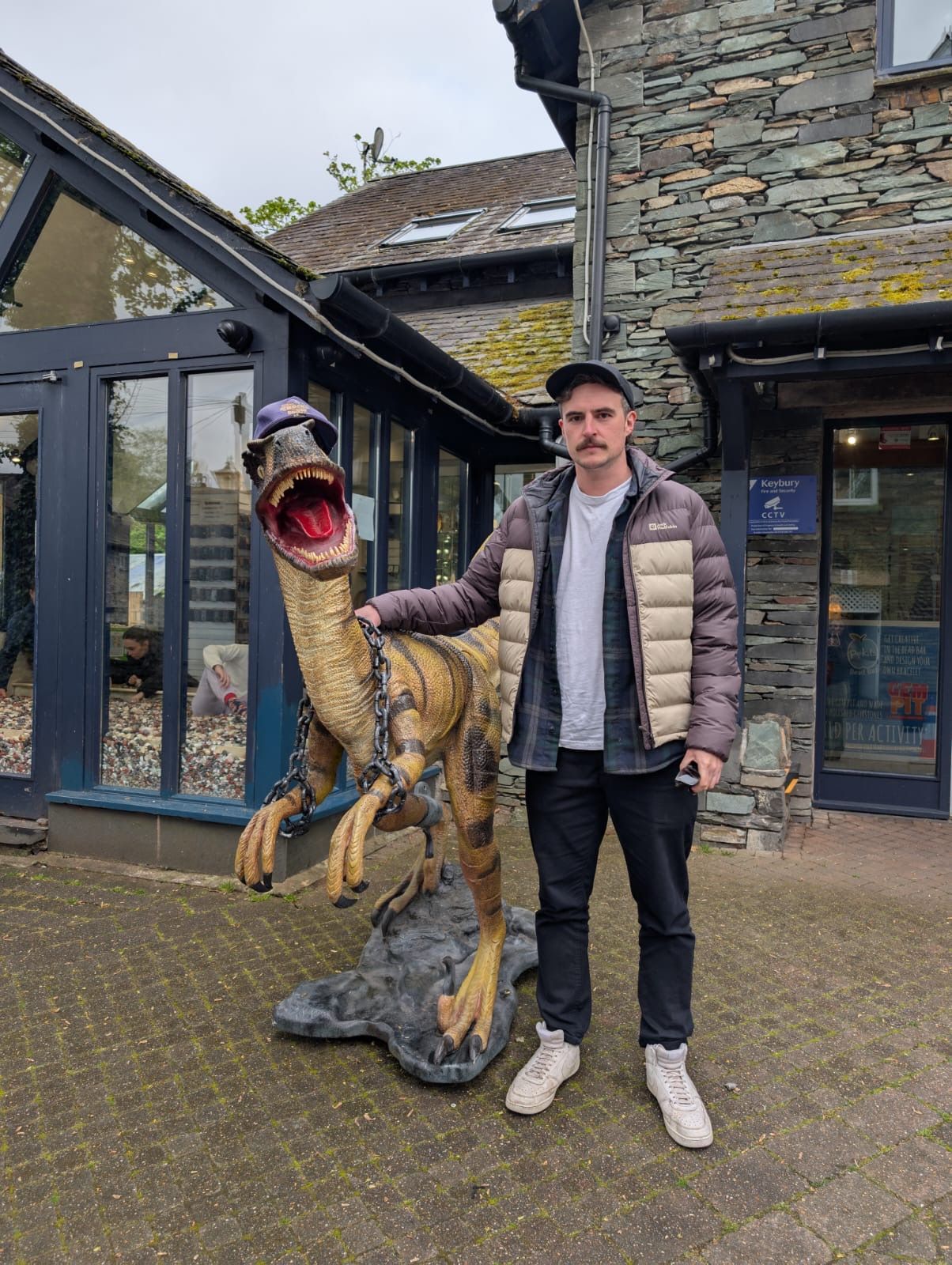 A man with a mustache poses beside a realistic velociraptor statue outside a stone building.