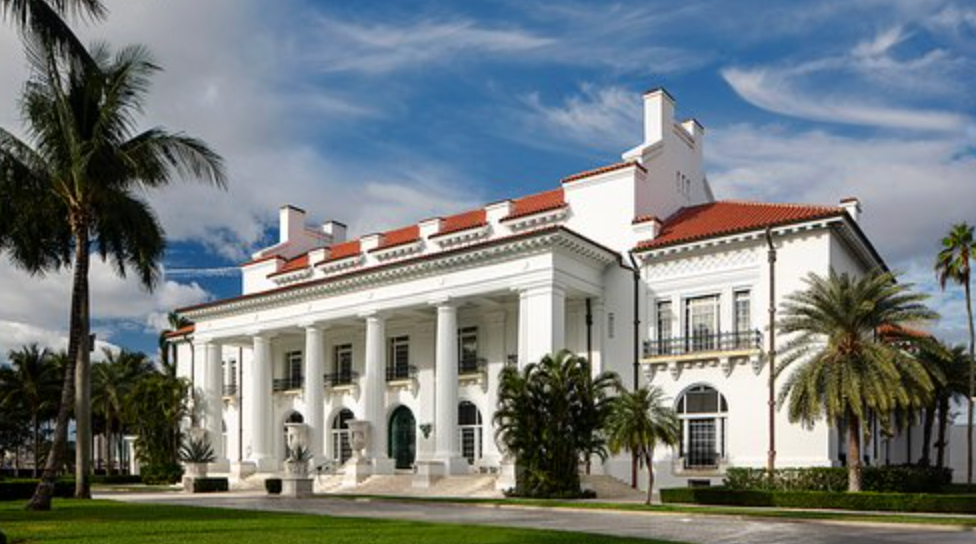 White Mediterranean-style mansion with red tile roof, grand columns, and lush palm trees under a partly cloudy blue sky.