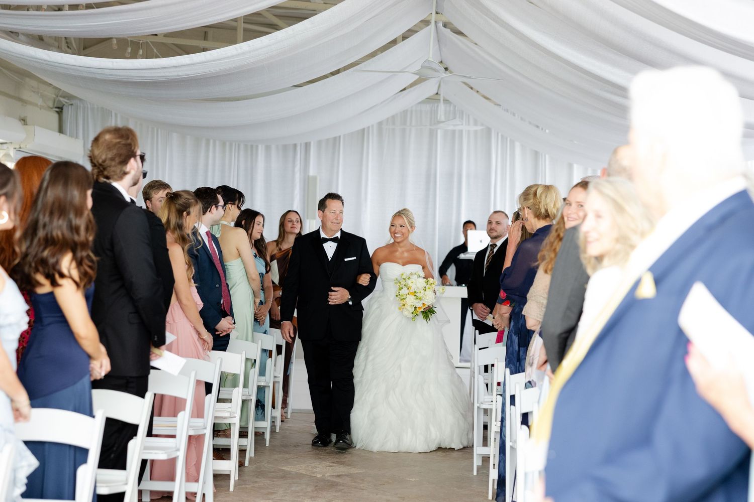Bride walks down the aisle in a white tent ceremony venue surrounded by seated guests in white folding chairs.