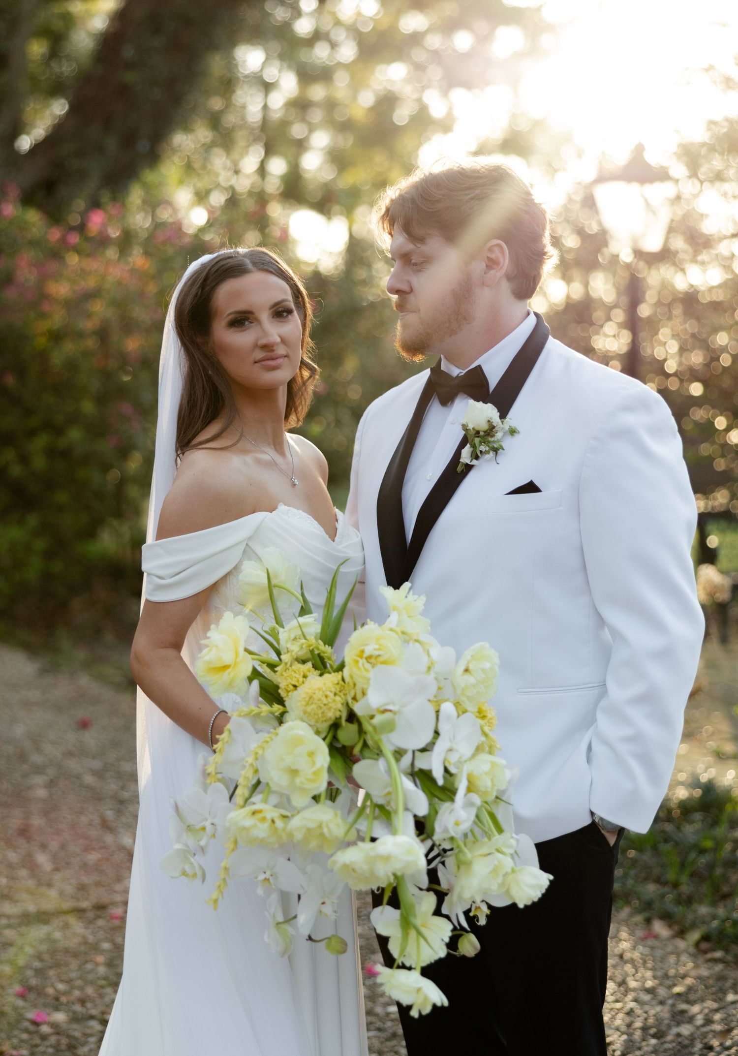 Bride and groom posing outdoors at golden hour, bride holding yellow and white cascading floral bouquet.