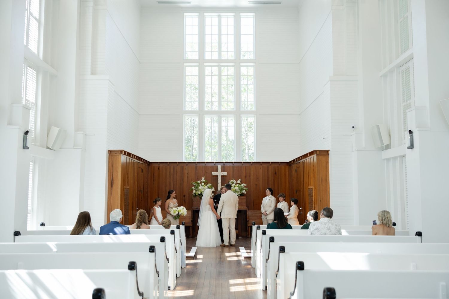 Elegant wedding ceremony in a bright white chapel with wooden pews, couple at altar beneath a cross and tall windows.