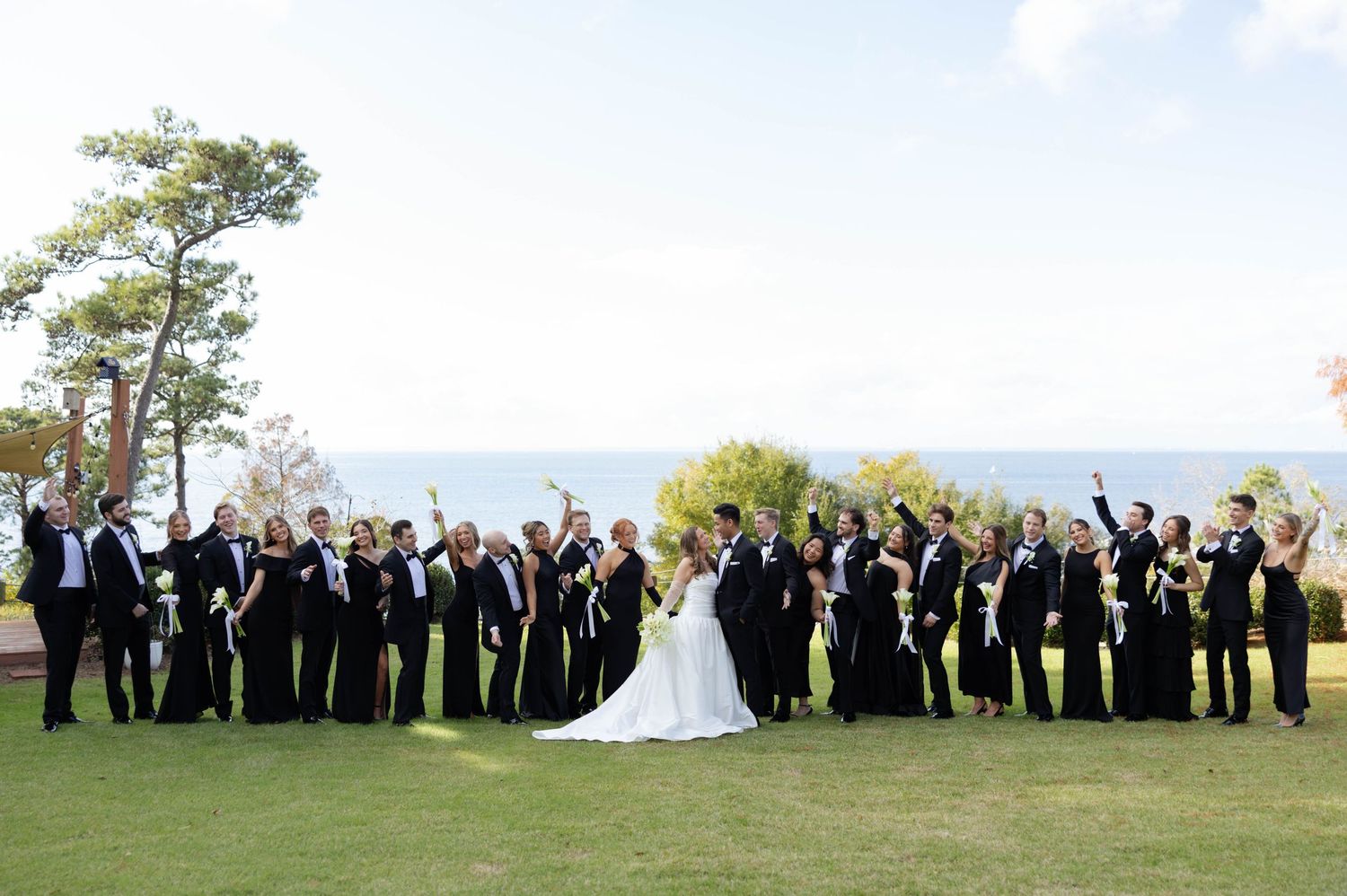 Large wedding party celebrates on a green lawn with ocean views, all dressed in black attire.