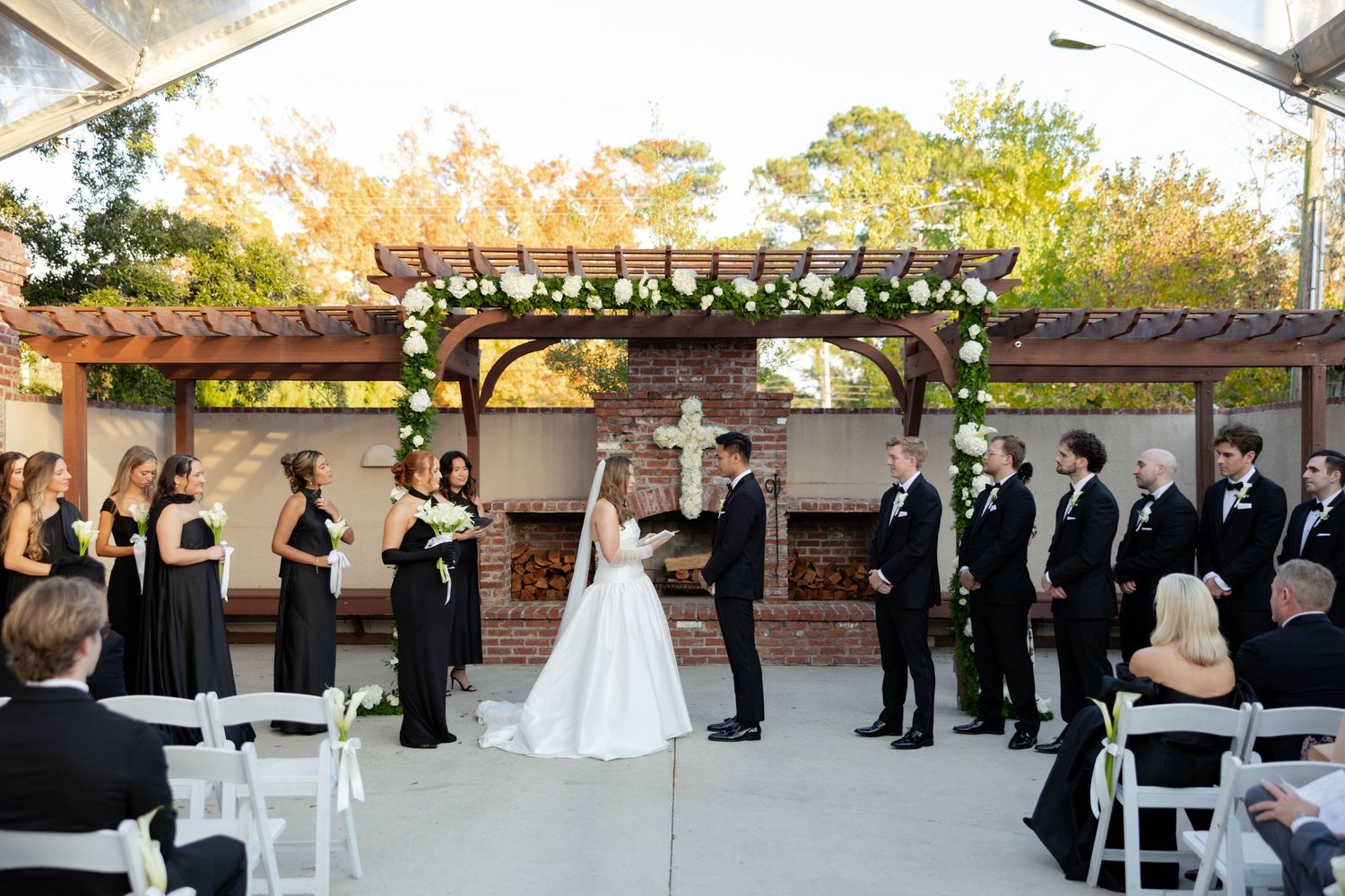 Outdoor wedding ceremony under floral arch with bridesmaids in black dresses and groomsmen beside rustic brick pergola.