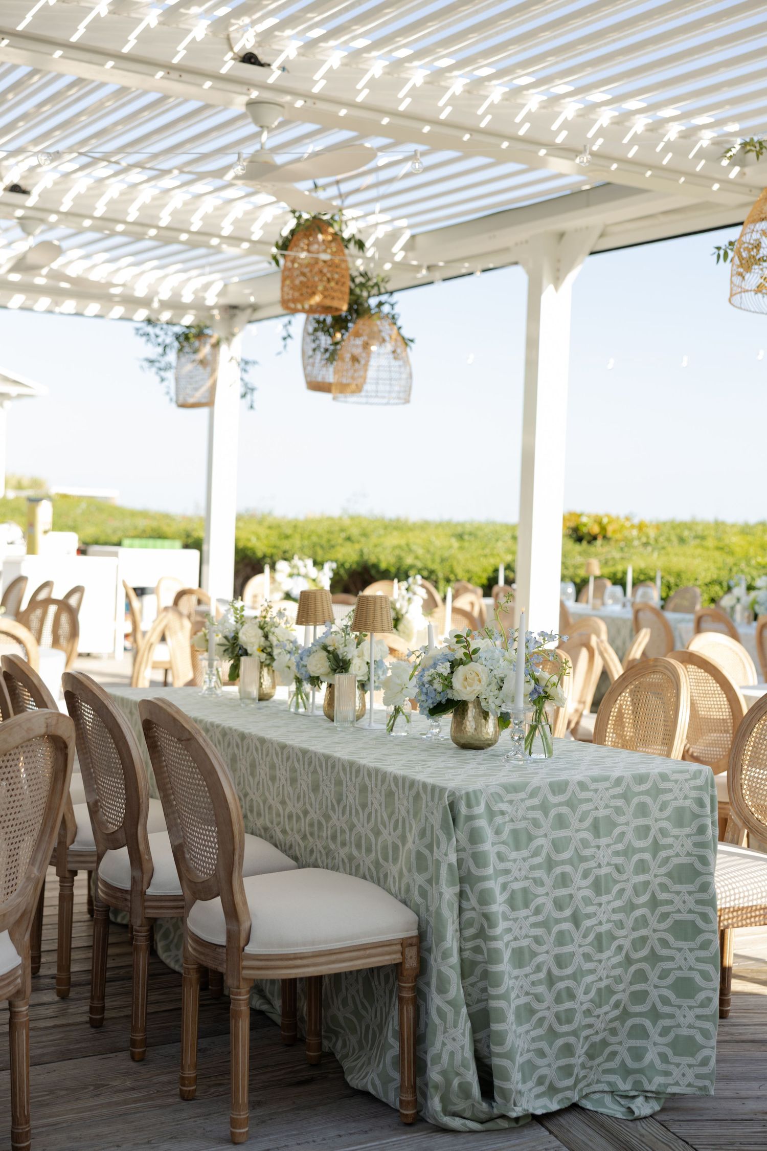 Elegant outdoor wedding reception with sage green patterned tablecloth, white floral centerpieces, and rattan chairs under a pergola.