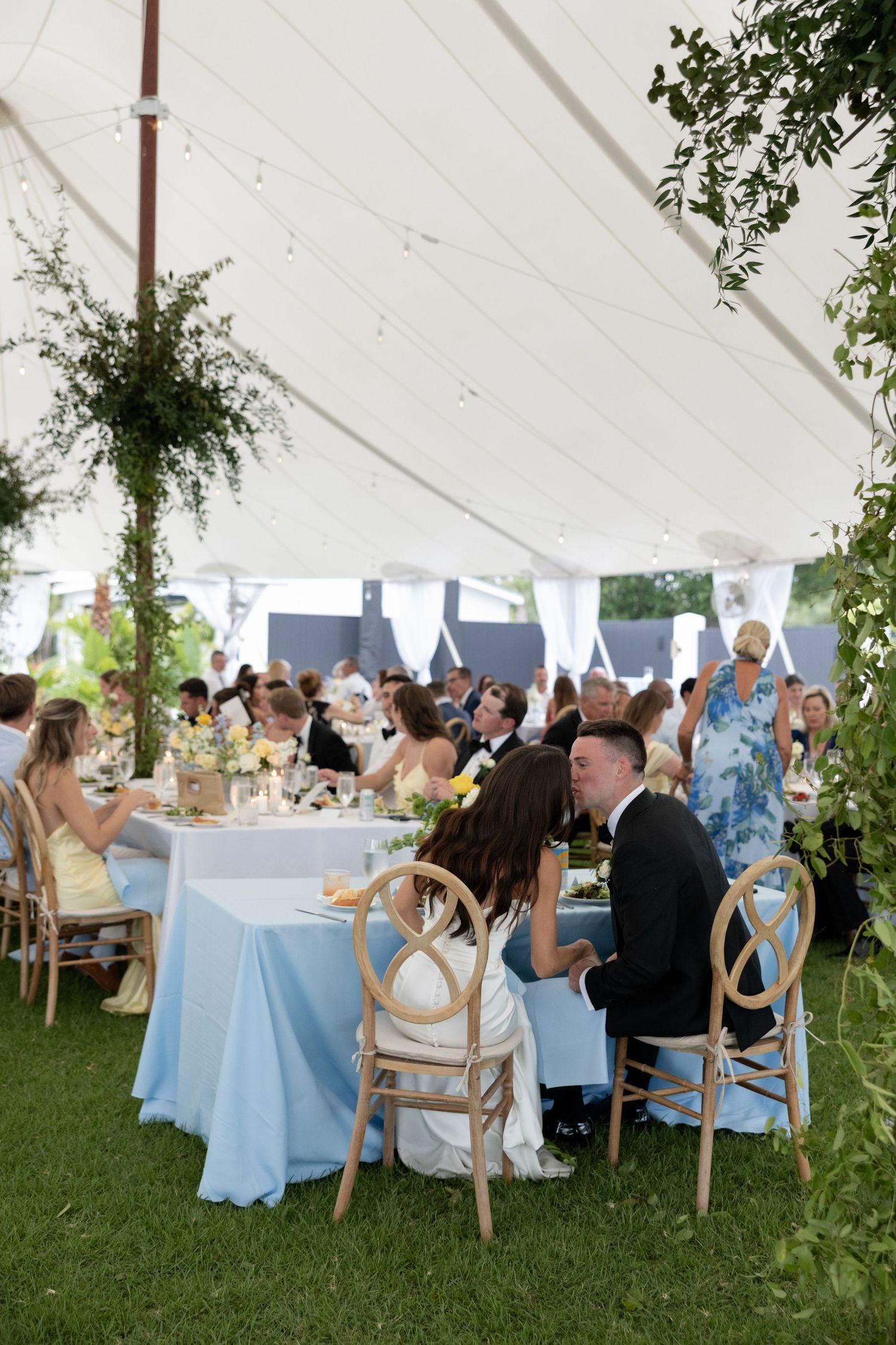 Elegant outdoor wedding reception under a white tent with blue tablecloths, floral decor, and guests seated at round tables.