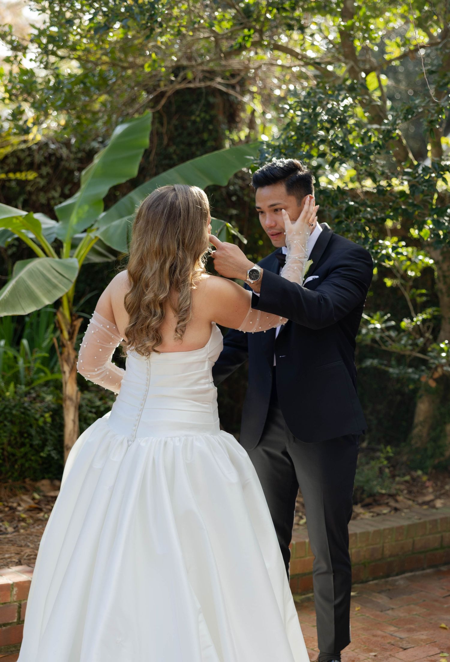Emotional groom wipes tears as bride approaches during a heartfelt first look moment in a lush garden setting.