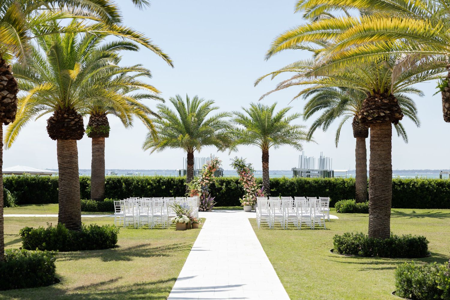 Outdoor wedding ceremony setup with white chairs, floral arch, palm trees, and ocean view on a sunny day.