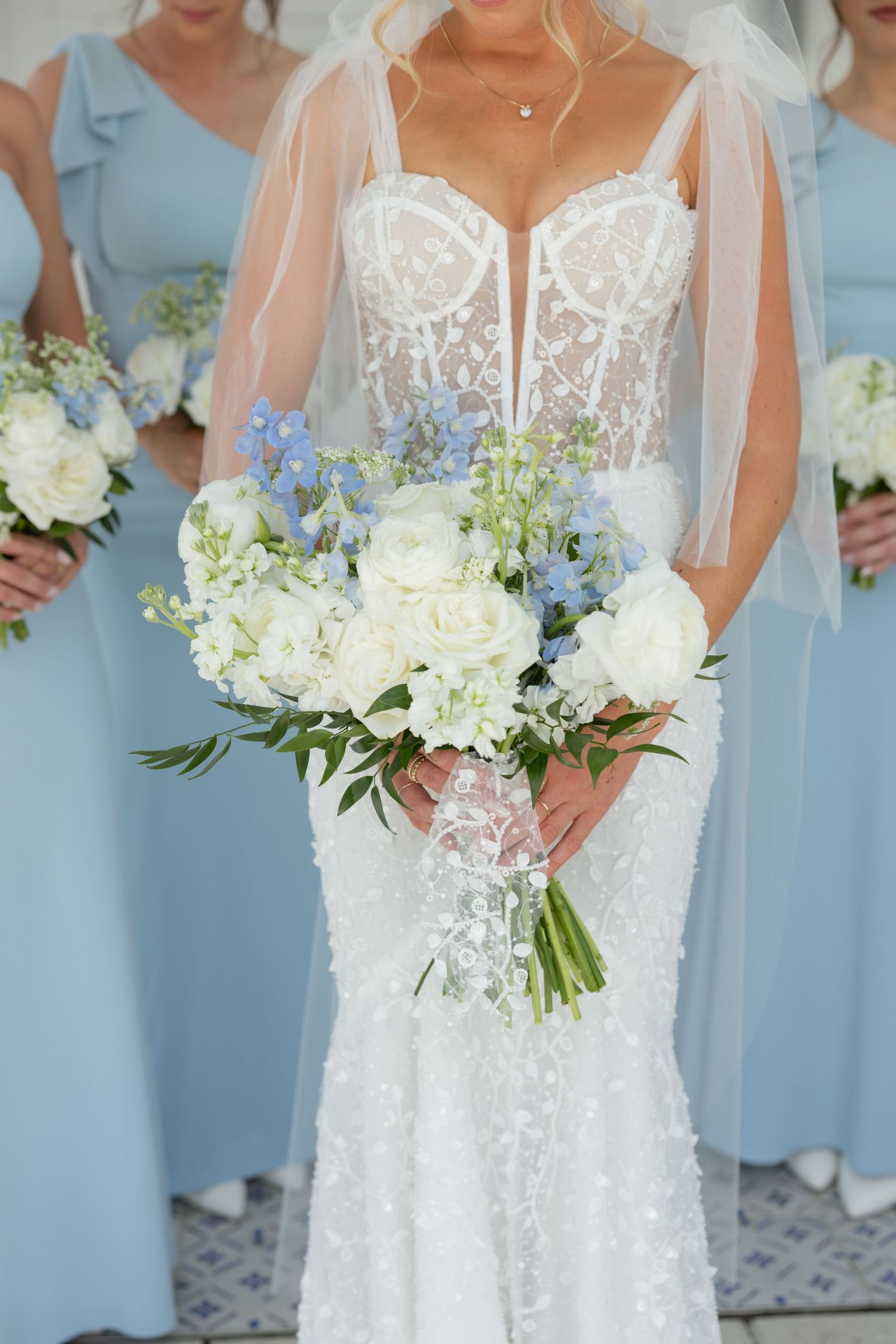 Bride in a lace mermaid gown holds a stunning white rose and blue delphinium bouquet with bridesmaids.